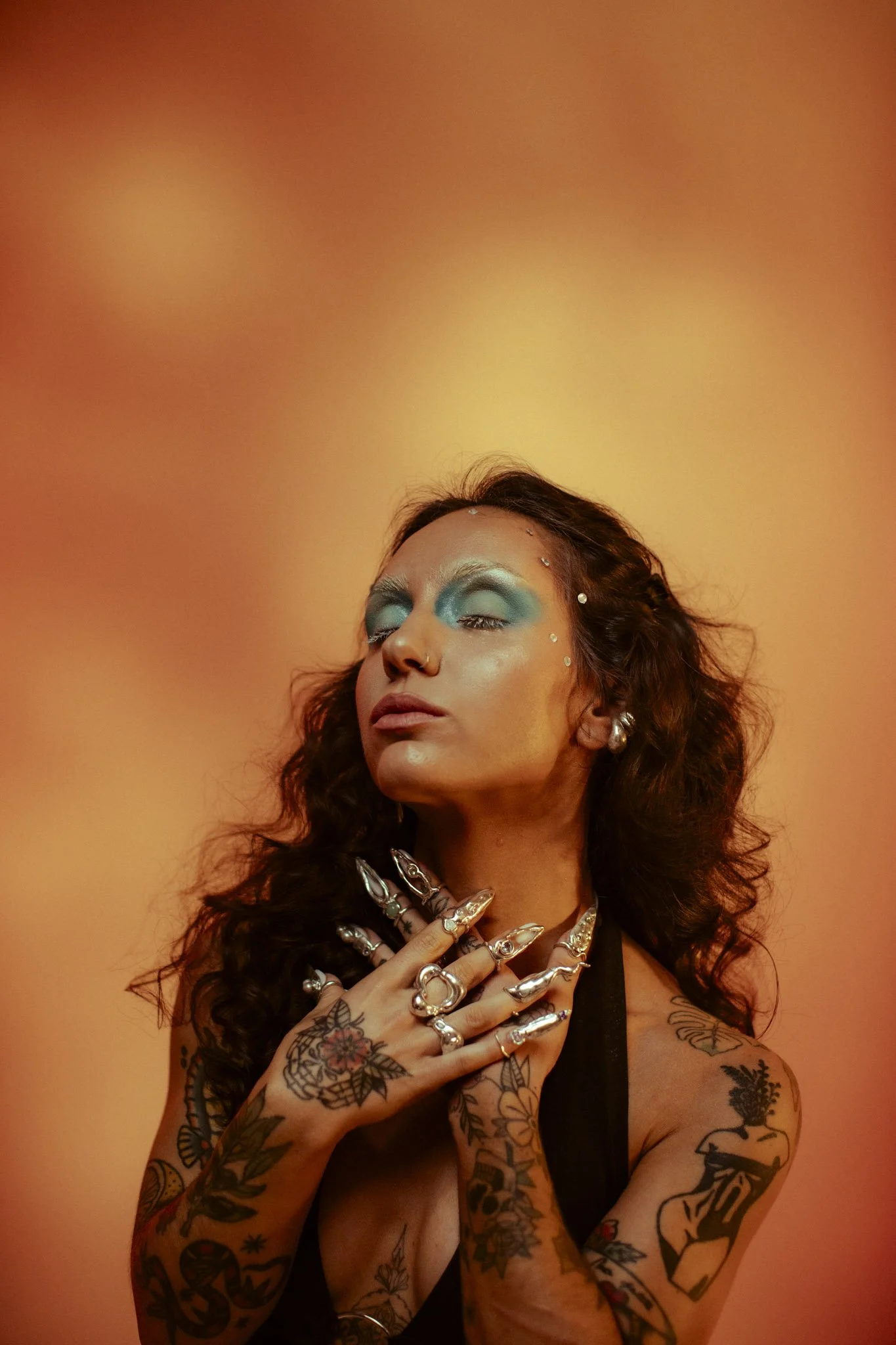 A woman with long curly hair, wearing blue eyeshadow and multiple silver rings and earrings, is posing with her eyes closed and her hands over her chest, showcasing tattoos on her arms and hands, against an orange background.