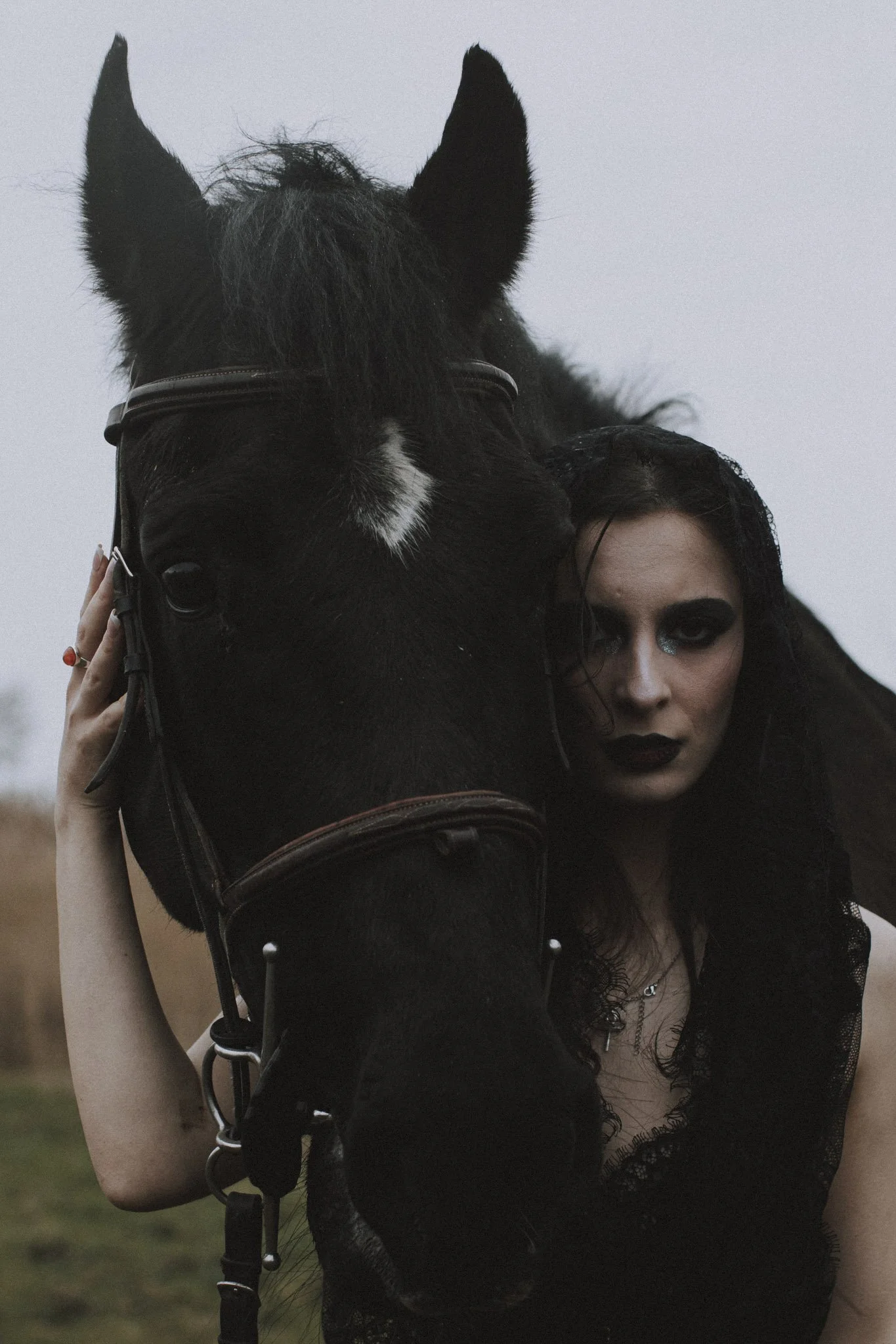 A person with dark curly hair is partly visible, holding the bridle of a black horse with a white star on its forehead. The person has dark makeup and is wearing a black lace top. The background is blurred with a cloudy sky.