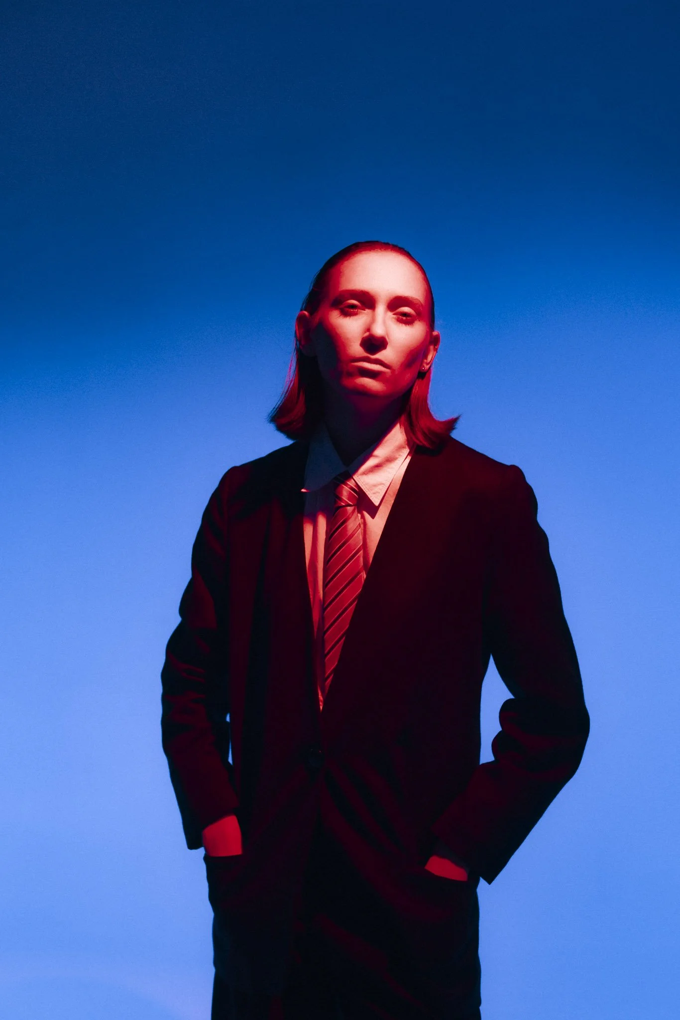 A person with long hair, wearing a black suit and striped tie, is standing with a gradient blue background, illuminated with red lighting on their face.