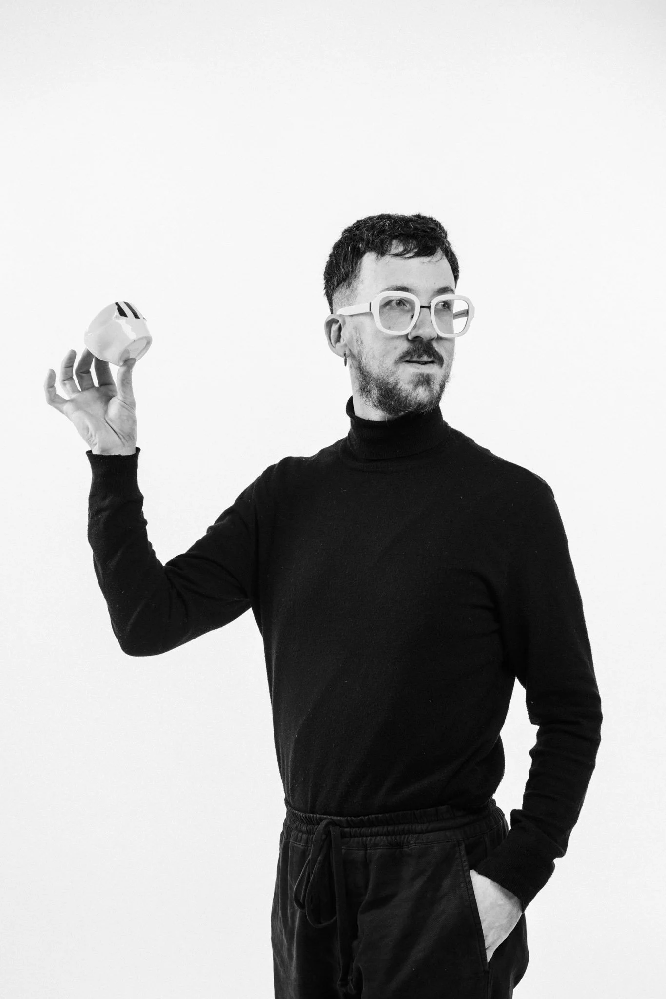 A man with short dark hair, a beard, and large white glasses, wearing a black turtleneck and black pants, is holding a small white ceramic object with black stripes in his right hand, against a plain white background.