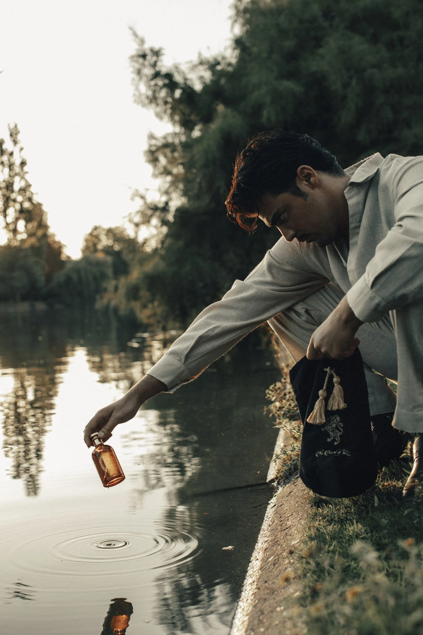 A man with dark hair, wearing a beige shirt, is sitting by a riverbank during sunset, holding a lantern in his right hand and releasing a bottle into the water with his left hand.