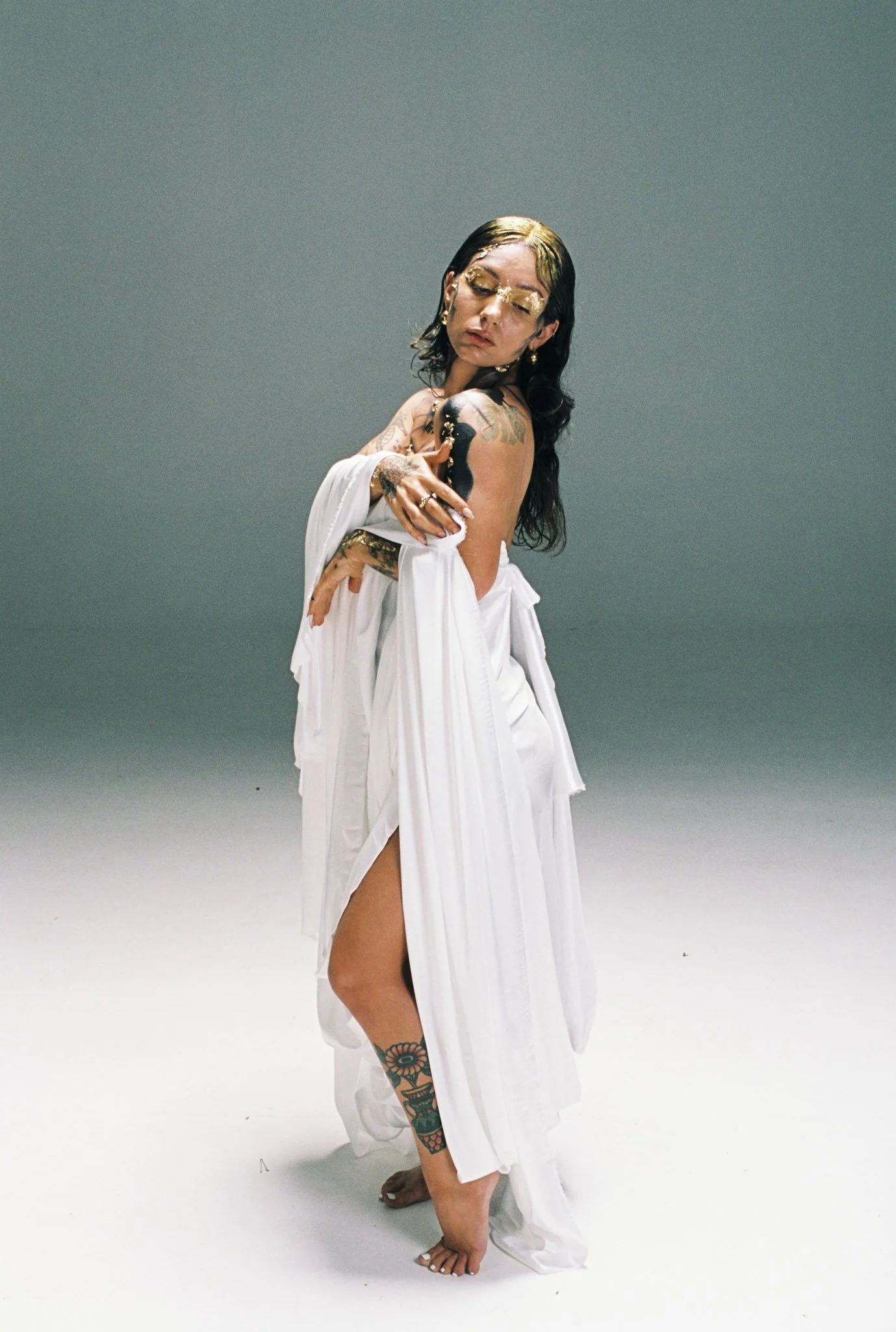 A woman with long dark hair and tattoos on her arms and leg, wearing a white draped dress, standing barefoot against a plain, muted background with her eyes closed and arms crossed over her chest.