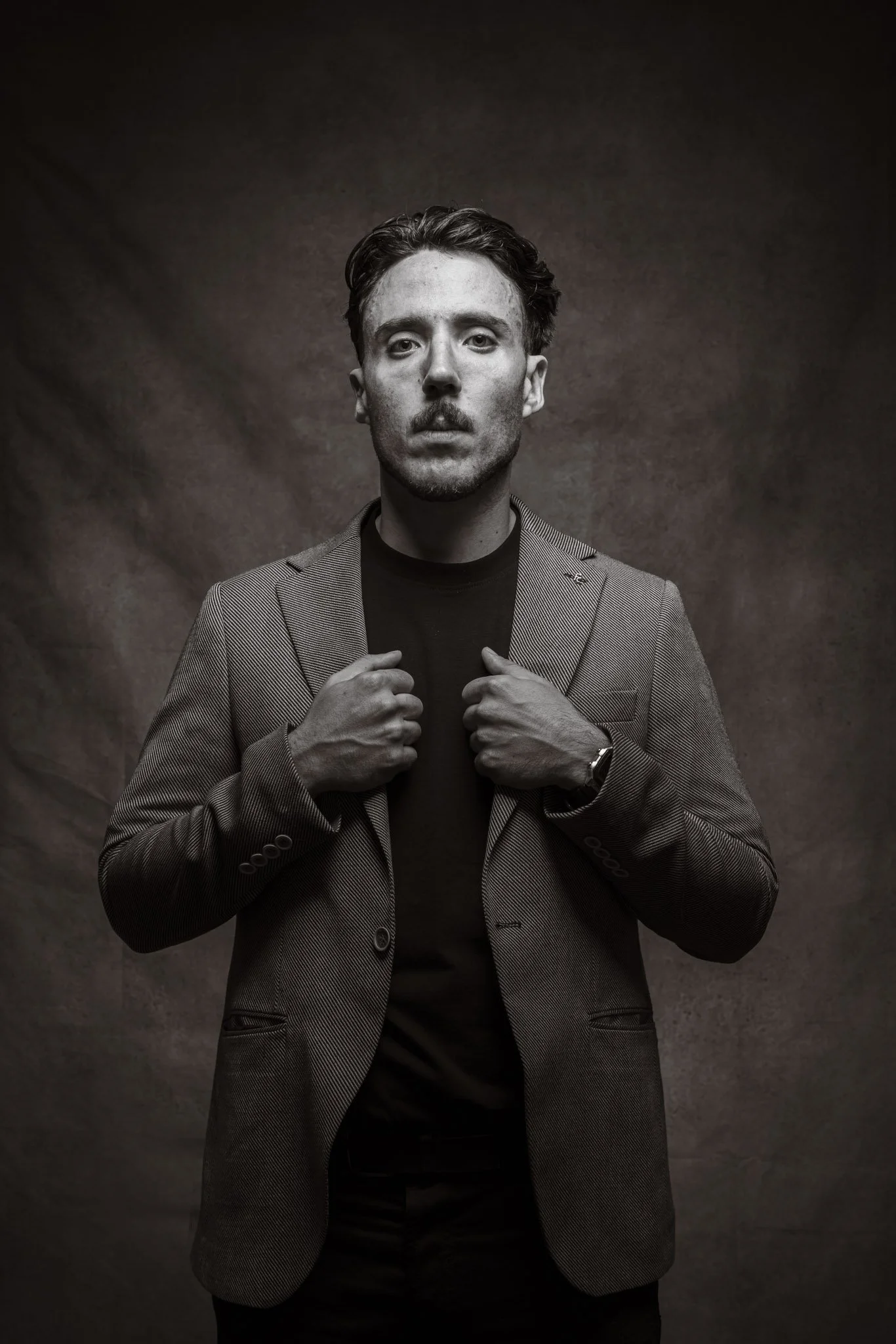 Black and white portrait of a man with no facial hair, wearing a checked blazer over a black shirt, standing against a textured dark background, holding the lapels of his blazer with both hands.