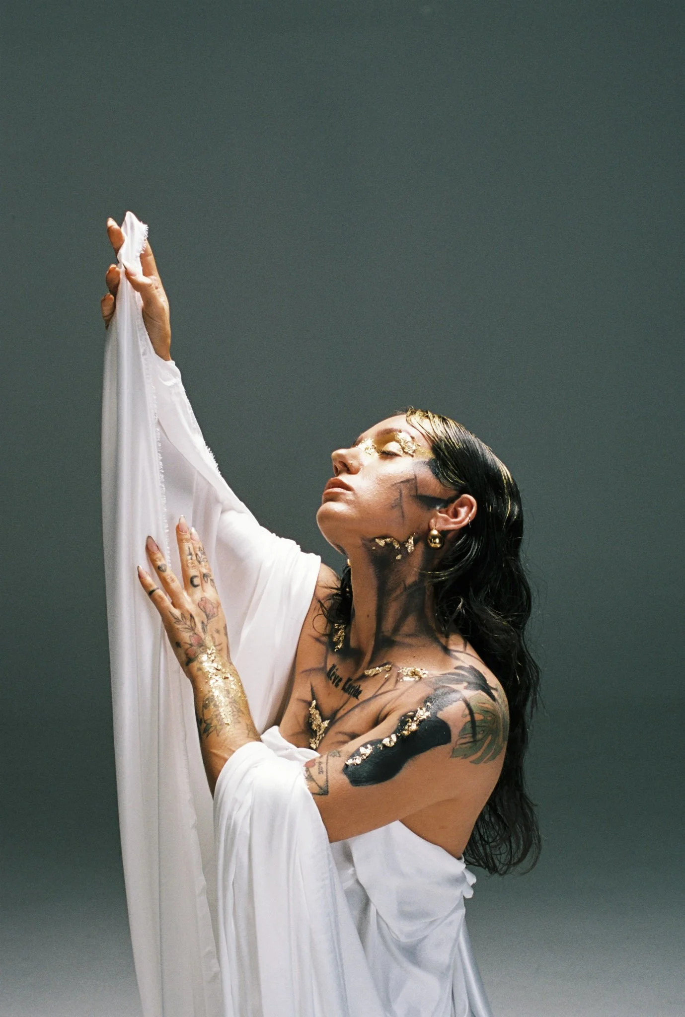 A woman with tattoos on her arms and face, wearing a white top, is holding a white cloth or garment with her right hand raised. Her eyes are closed, and she has gold accents and makeup on her face. The background is a plain, dark, muted color.