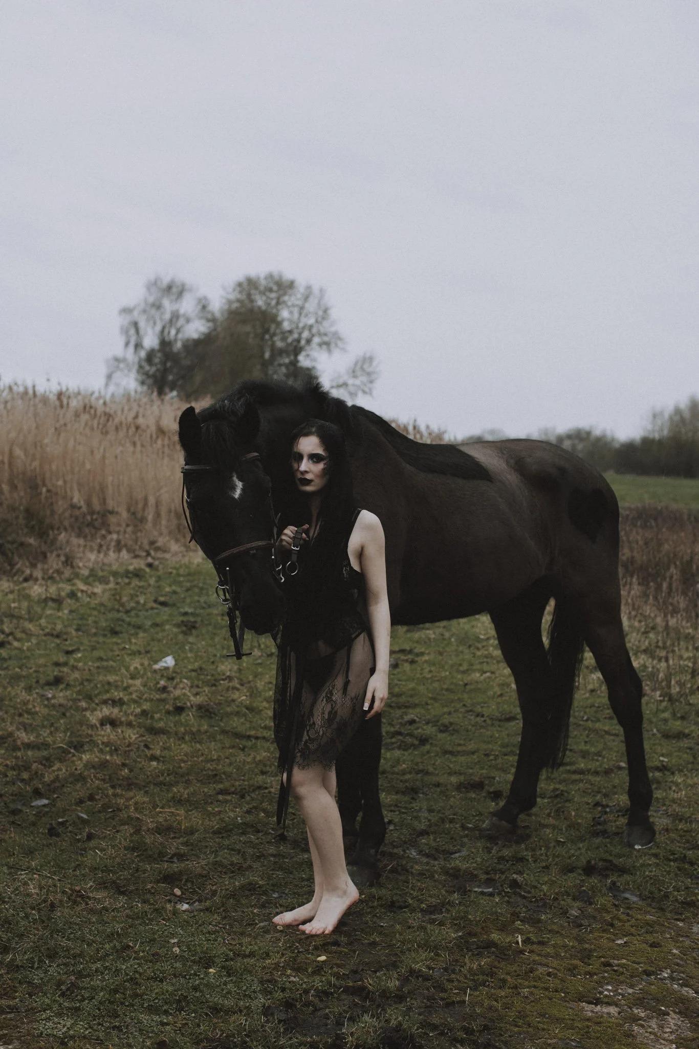 A person with long dark hair and pale skin standing outdoors with a dark horse, wearing a black lace dress and makeup, in a natural setting with trees and grass overcast sky.