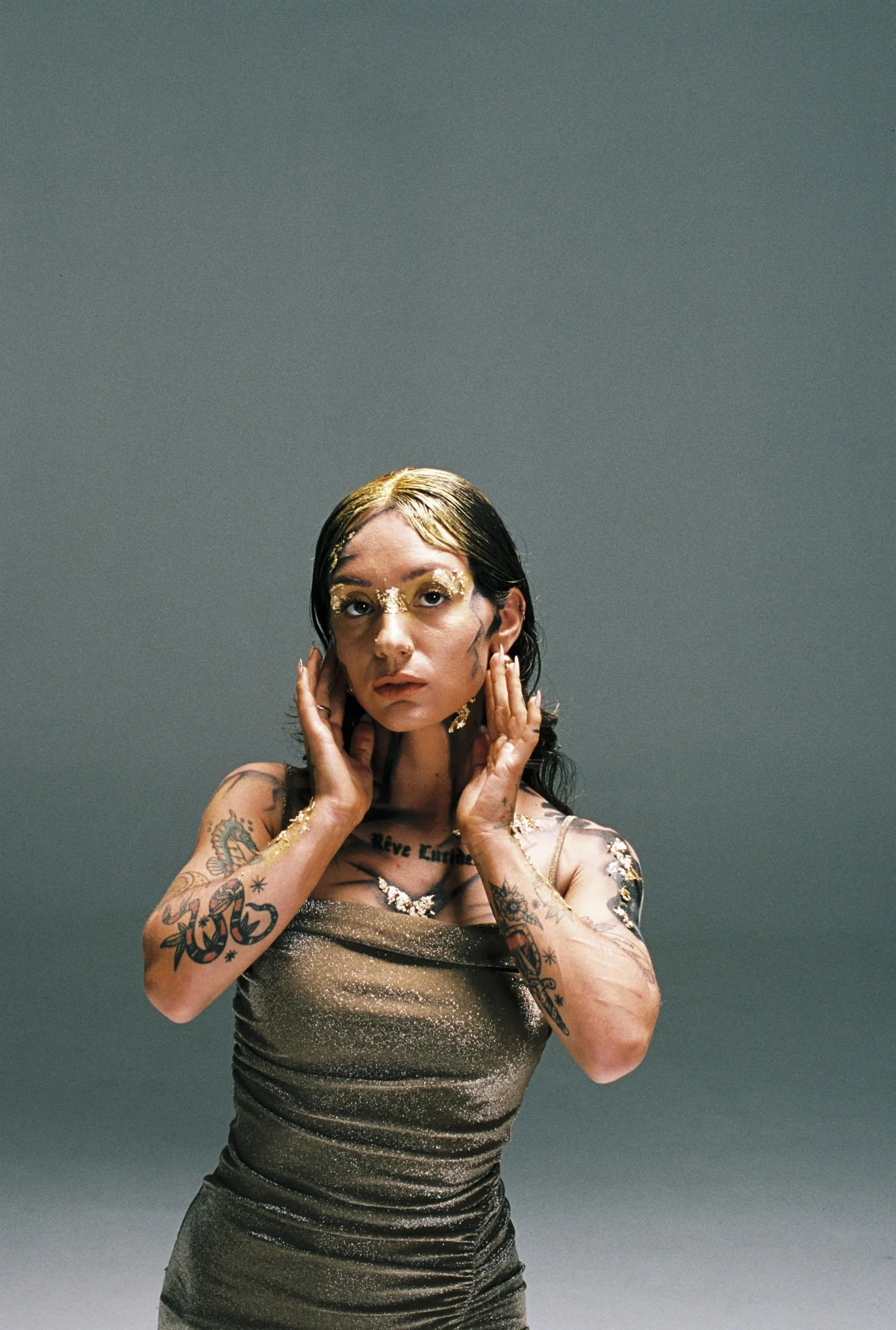A woman with tattoos and glitter makeup poses with her hands near her face against a plain background.