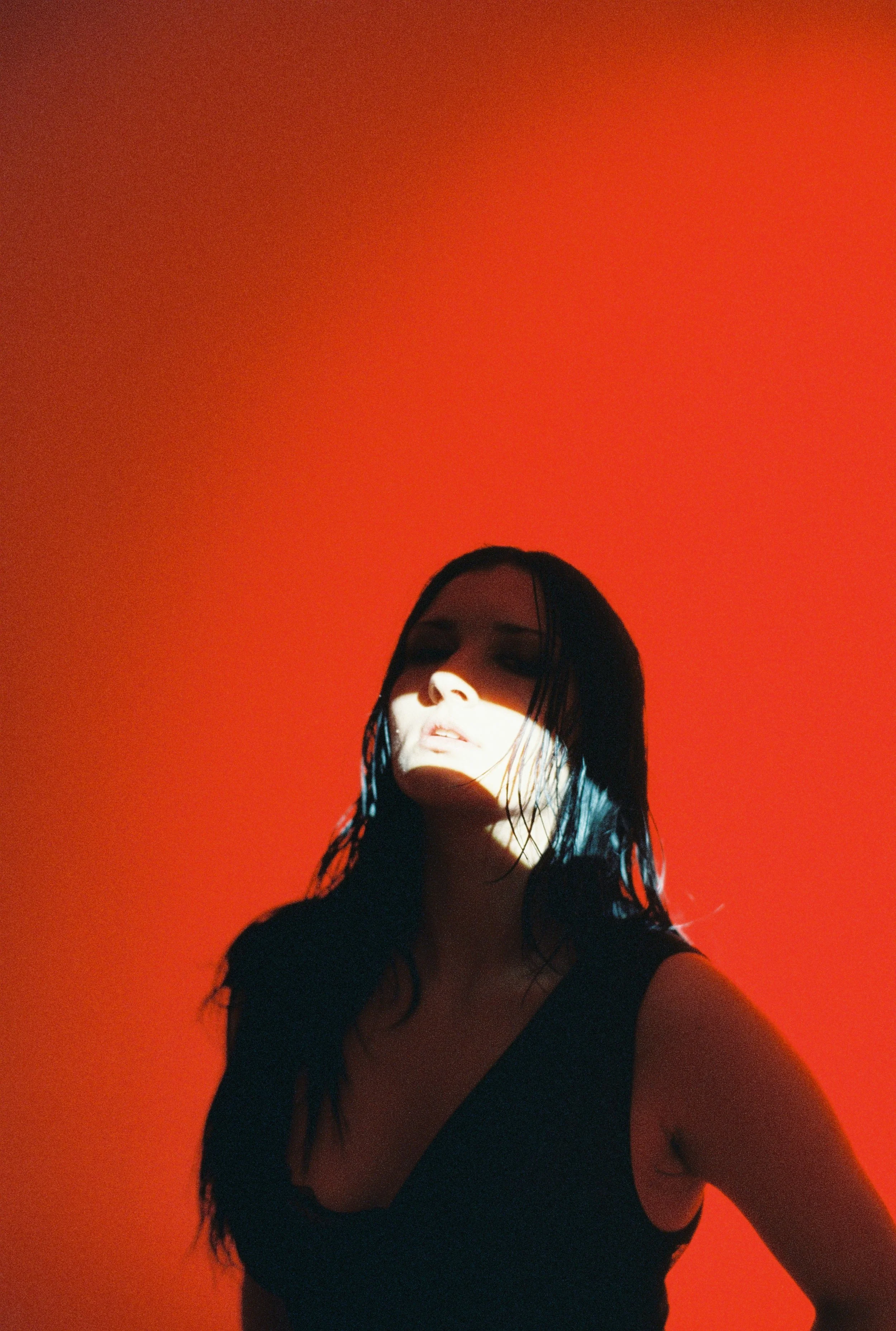 A woman with wet black hair is illuminated by a bright light, partially shadowed, against an orange background.