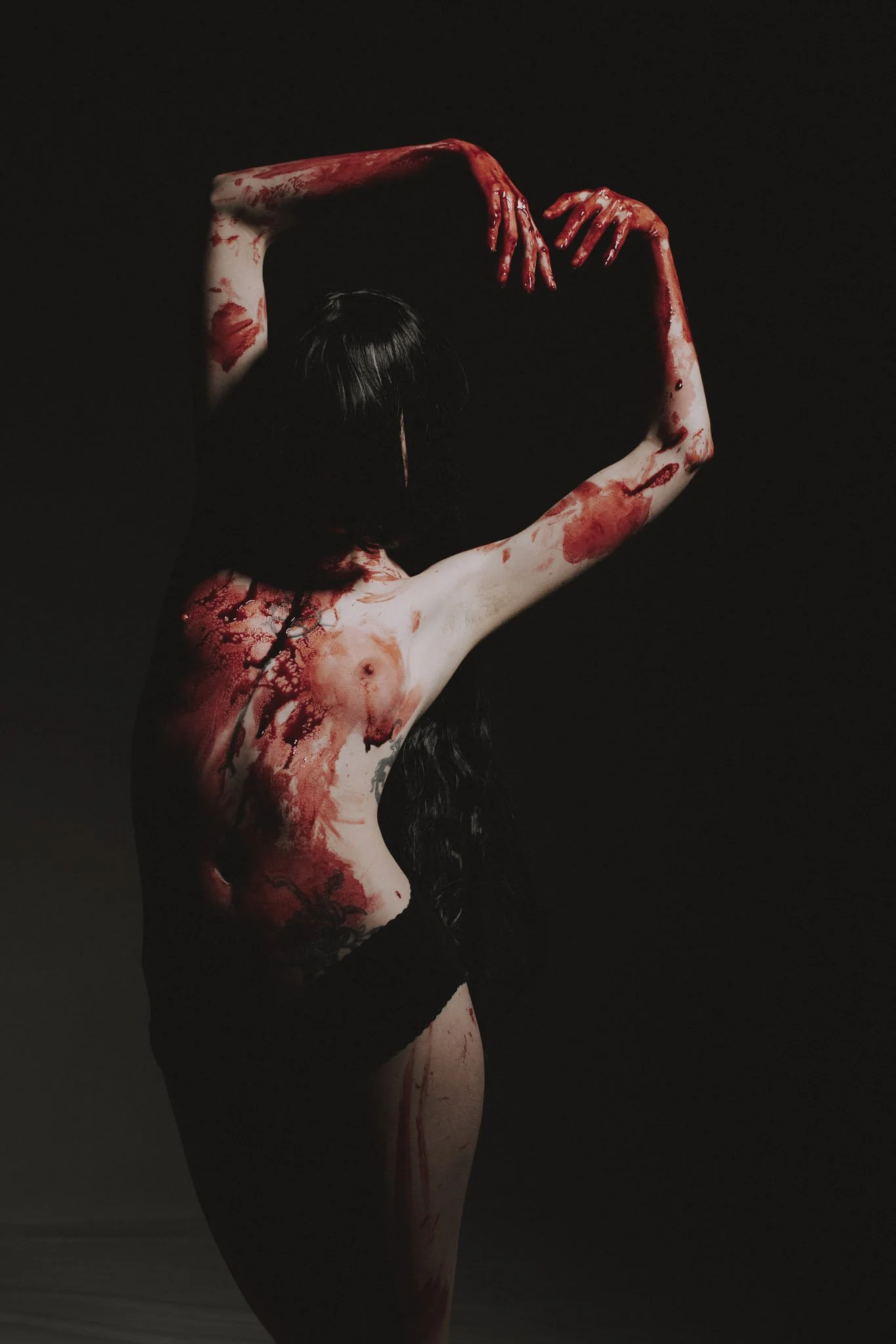 A woman with dark hair has her arms raised above her head, and her body and arms are covered with red paint or substance that looks like blood, set against a dark background.