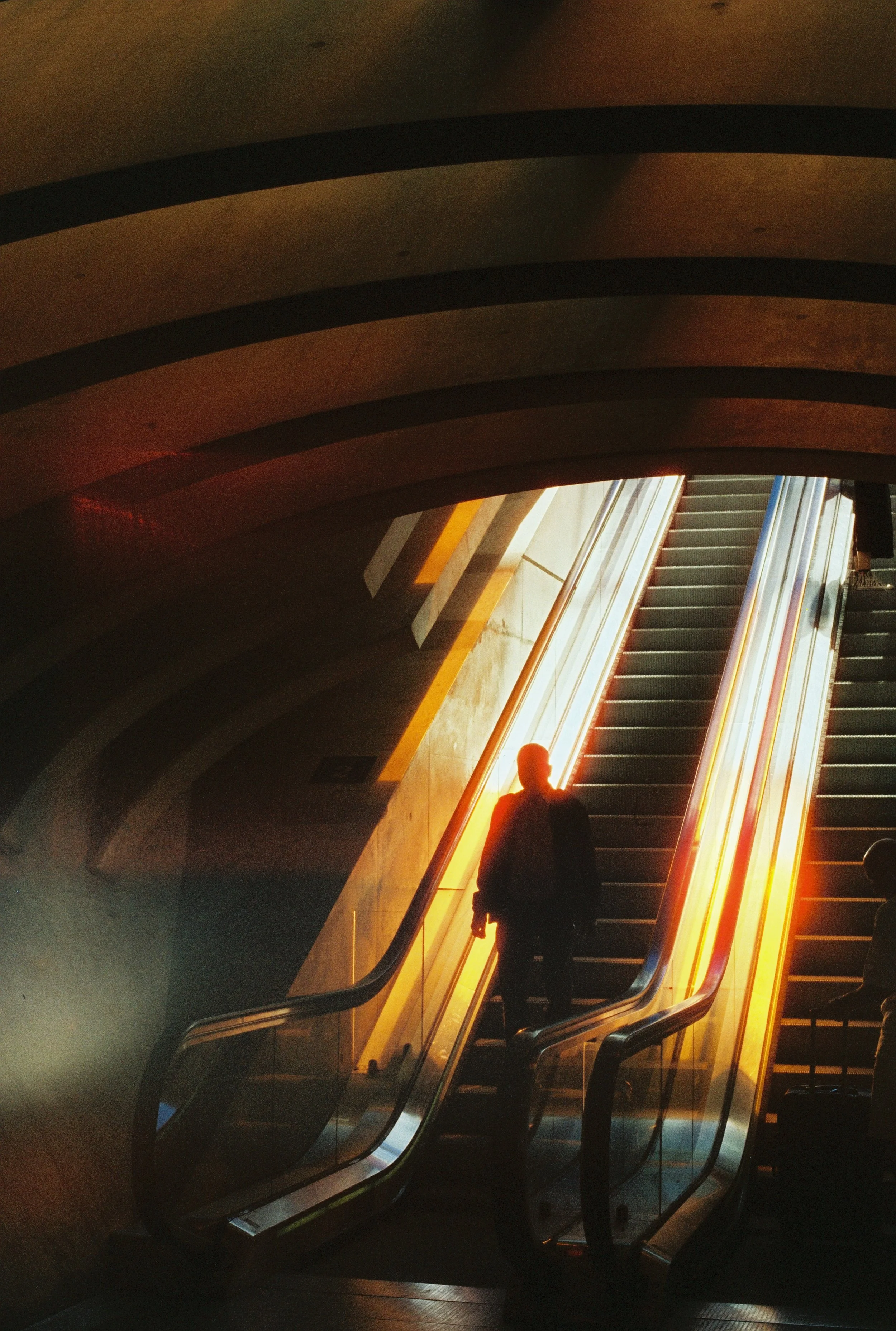 A person is walking down an escalator illuminated by warm, orange lighting. The escalator is located in a modern, indoor setting with curved architectural elements overhead.