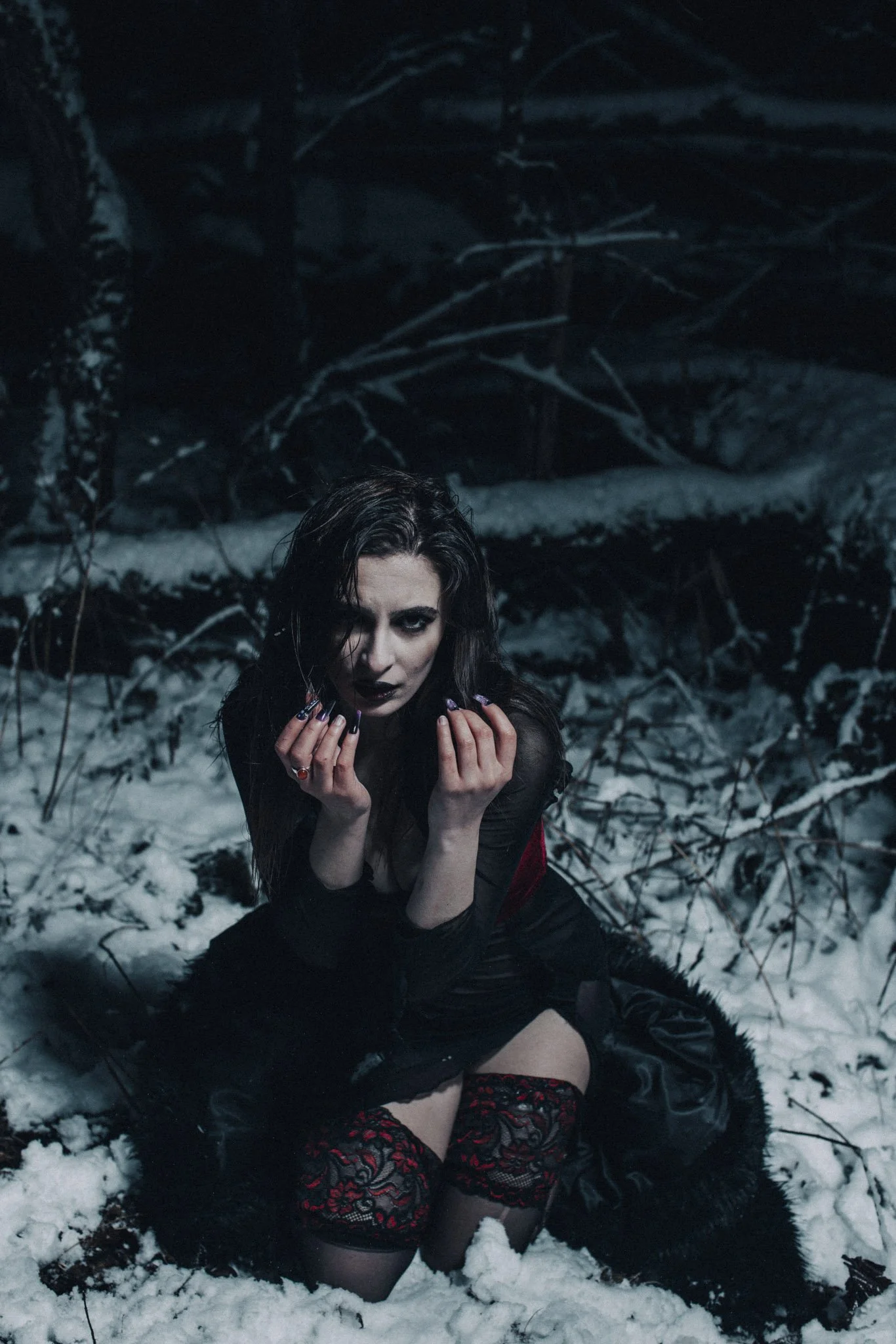 A woman with dark hair and makeup posing in a snowy forest at night, wearing black lace stockings, black gloves, and red accents, with dark clothing and a black fur cape.