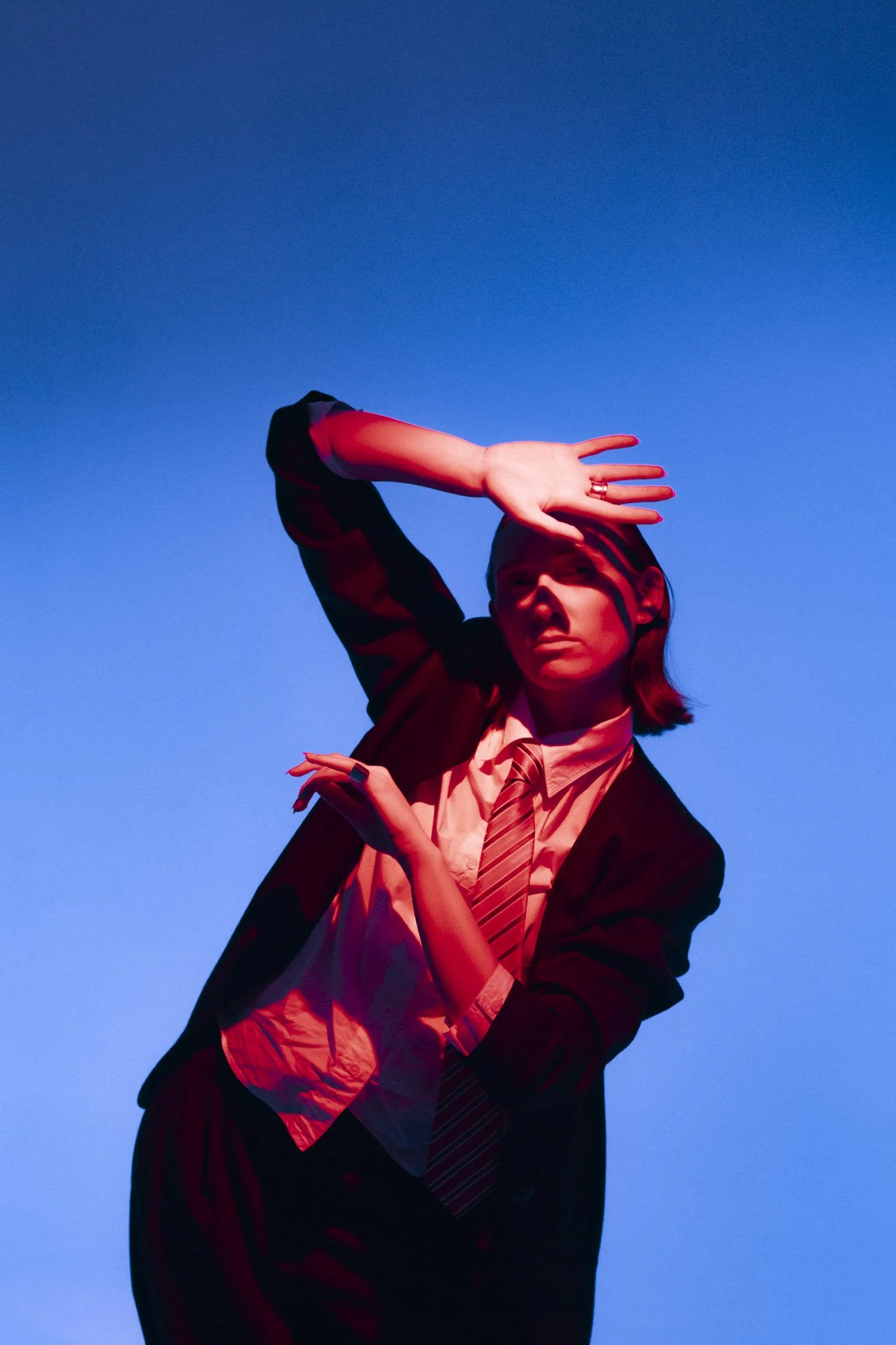 A woman posing against a blue sky, wearing a dark blazer, a white shirt, and a striped tie. She is holding her arm above her forehead and looking into the distance.