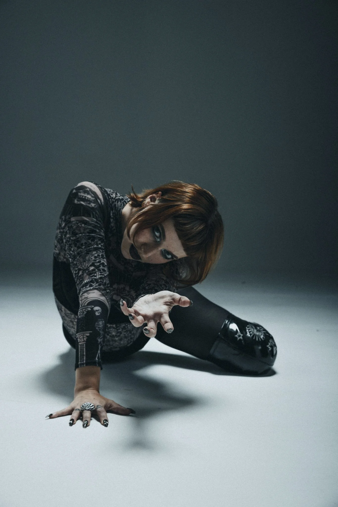 A woman with red hair and blue eyes, wearing black and white patterned clothing, is in a crouched, dynamic pose on the floor against a plain background.