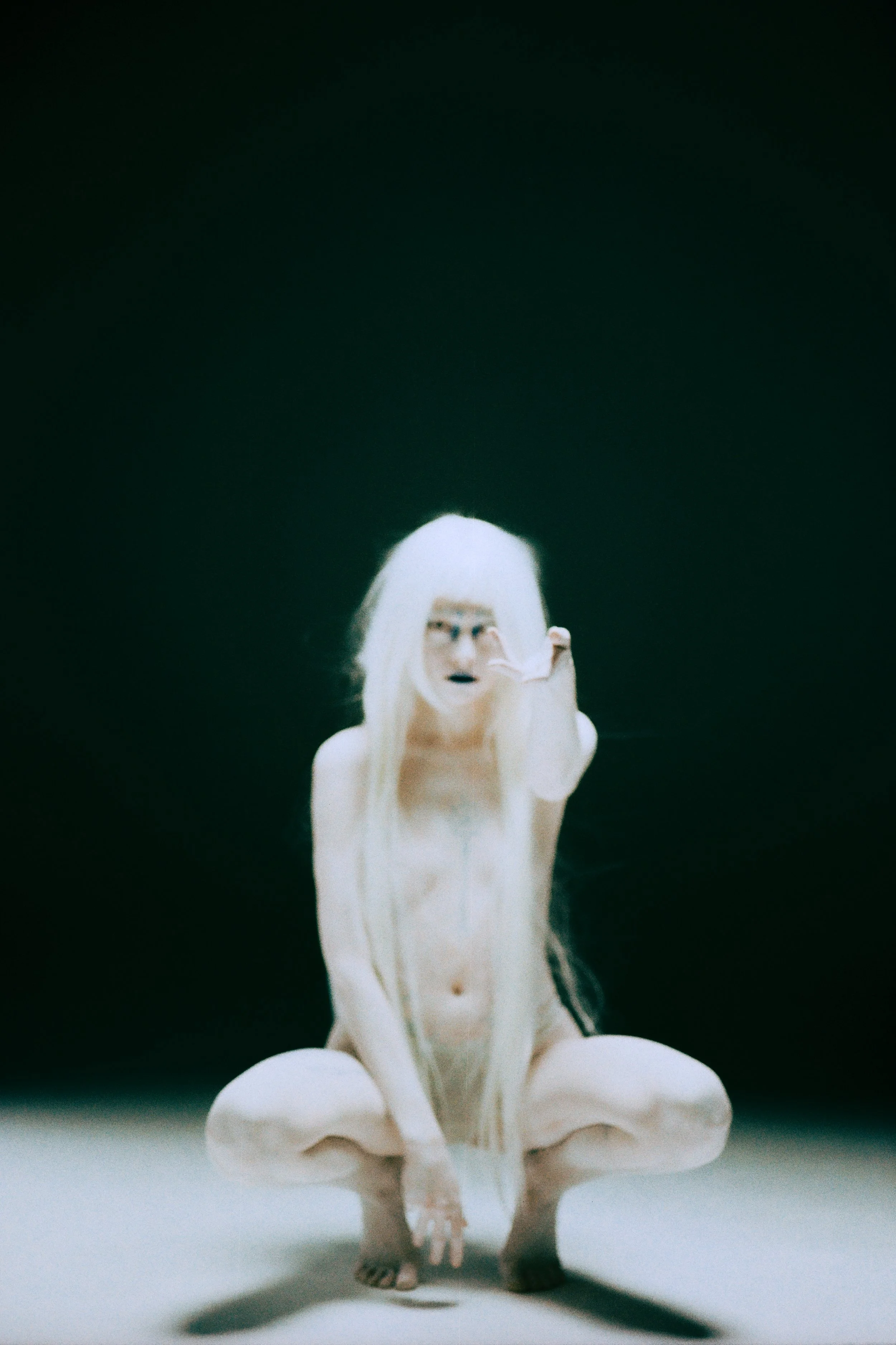 A person with long blonde hair wearing glasses and a white outfit, sitting on the ground and adjusting the glasses with one hand, against a dark background.