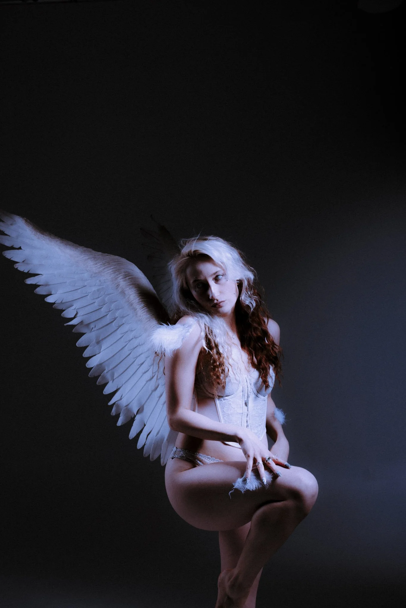 A woman with white feathered wings, long curly hair, and a white corset poses against a dark background.