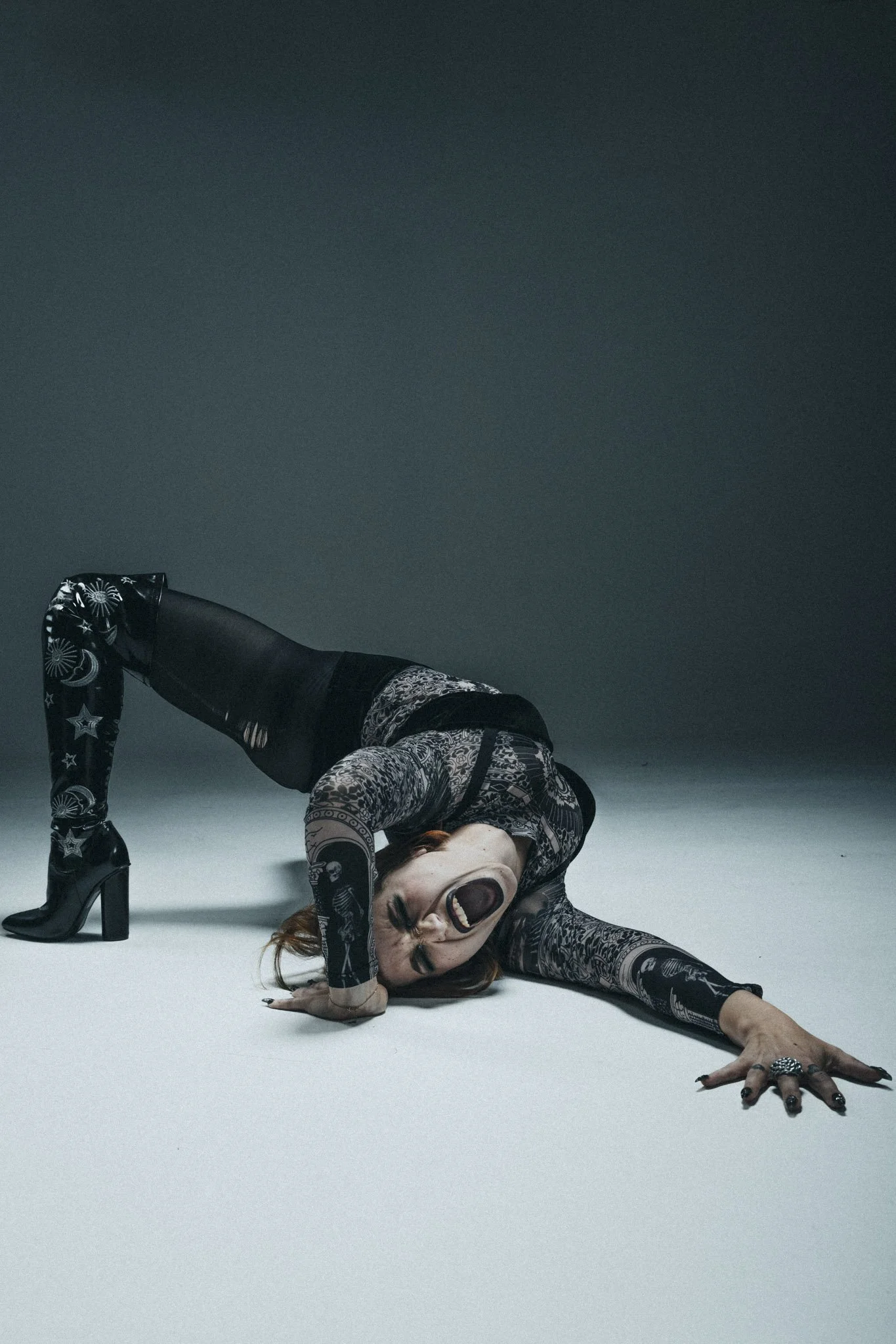 A woman in patterned clothing and high-heeled boots is on the floor in a pose with her head near the ground and an open mouth, wearing dark lipstick, rings, and a long sleeve top with skeleton print.