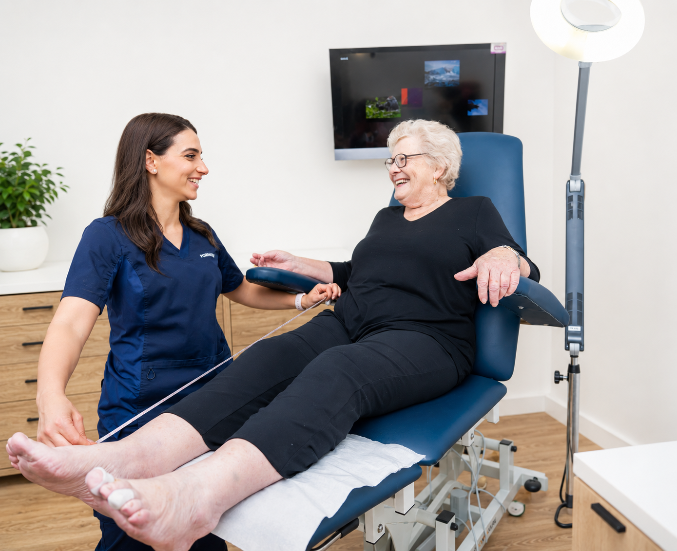 A healthcare professional, wearing navy scrubs, assisting an elderly woman with a medical examination in a clinic or hospital room. The elderly woman is seated on an exam table, smiling, with her legs extended and connected to monitoring equipment. A flat-screen monitor is on the wall in the background.