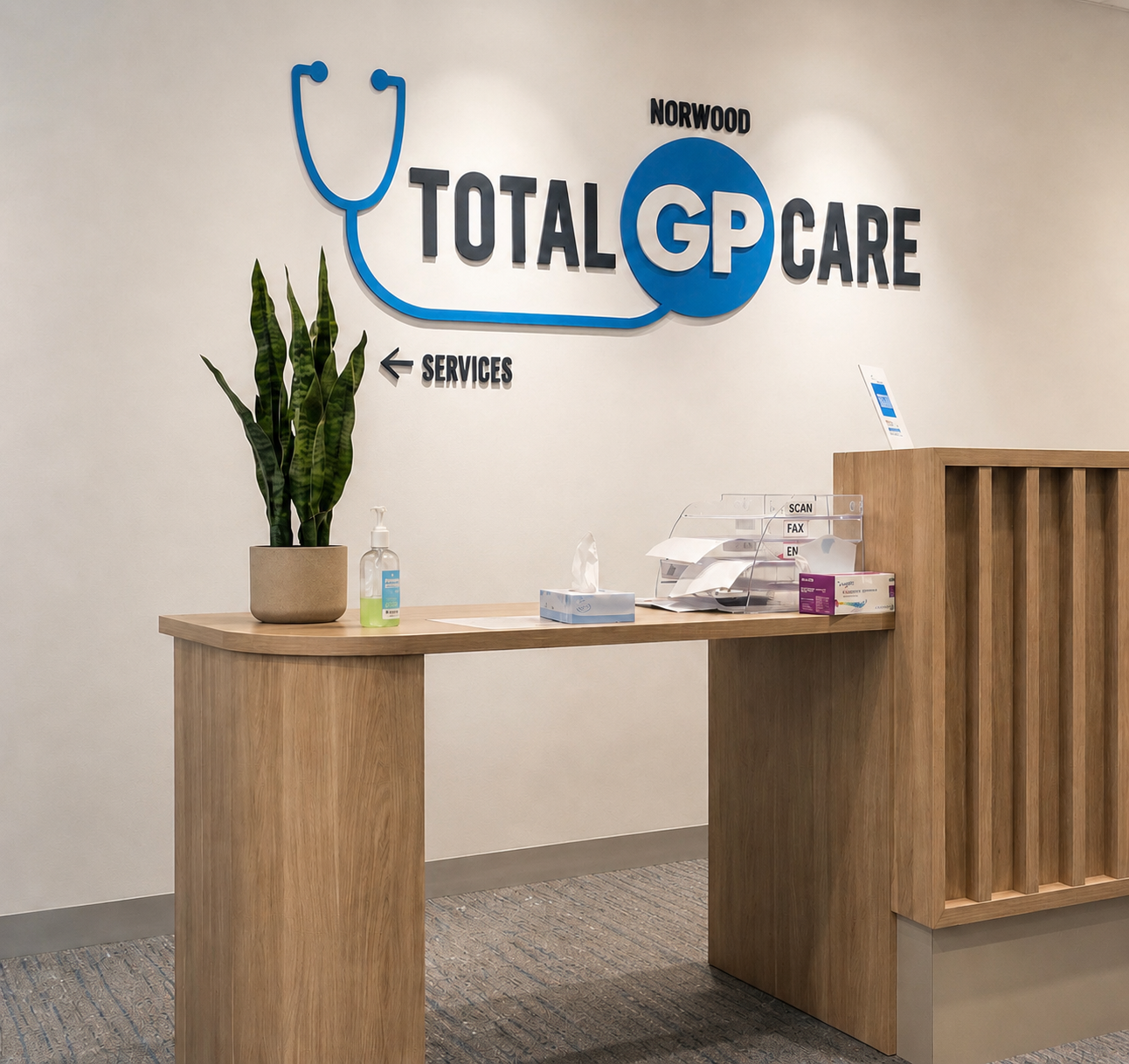Reception area of a healthcare clinic with a wooden desk, a green potted plant, hand sanitizer, and a clear paper organizer. The wall has a blue and black logo with a stethoscope shape and the text 'Total GP Care' and directional signs for services.