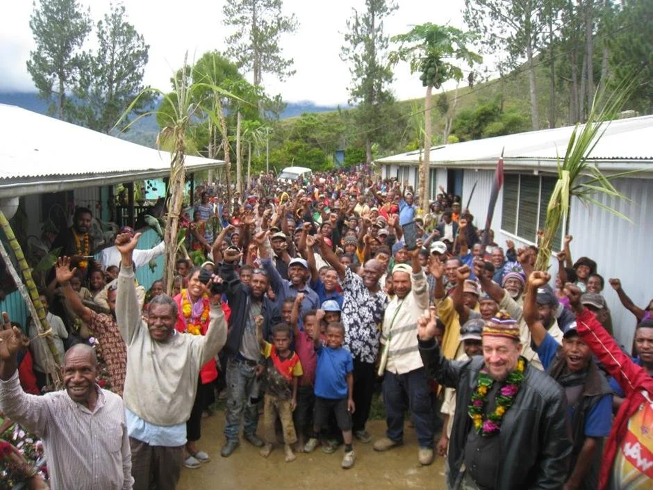 A large crowd of people gathered outdoors in a rural area, many holding palms and waving, with some wearing flower leis, and smiling or cheering during a celebration or event.