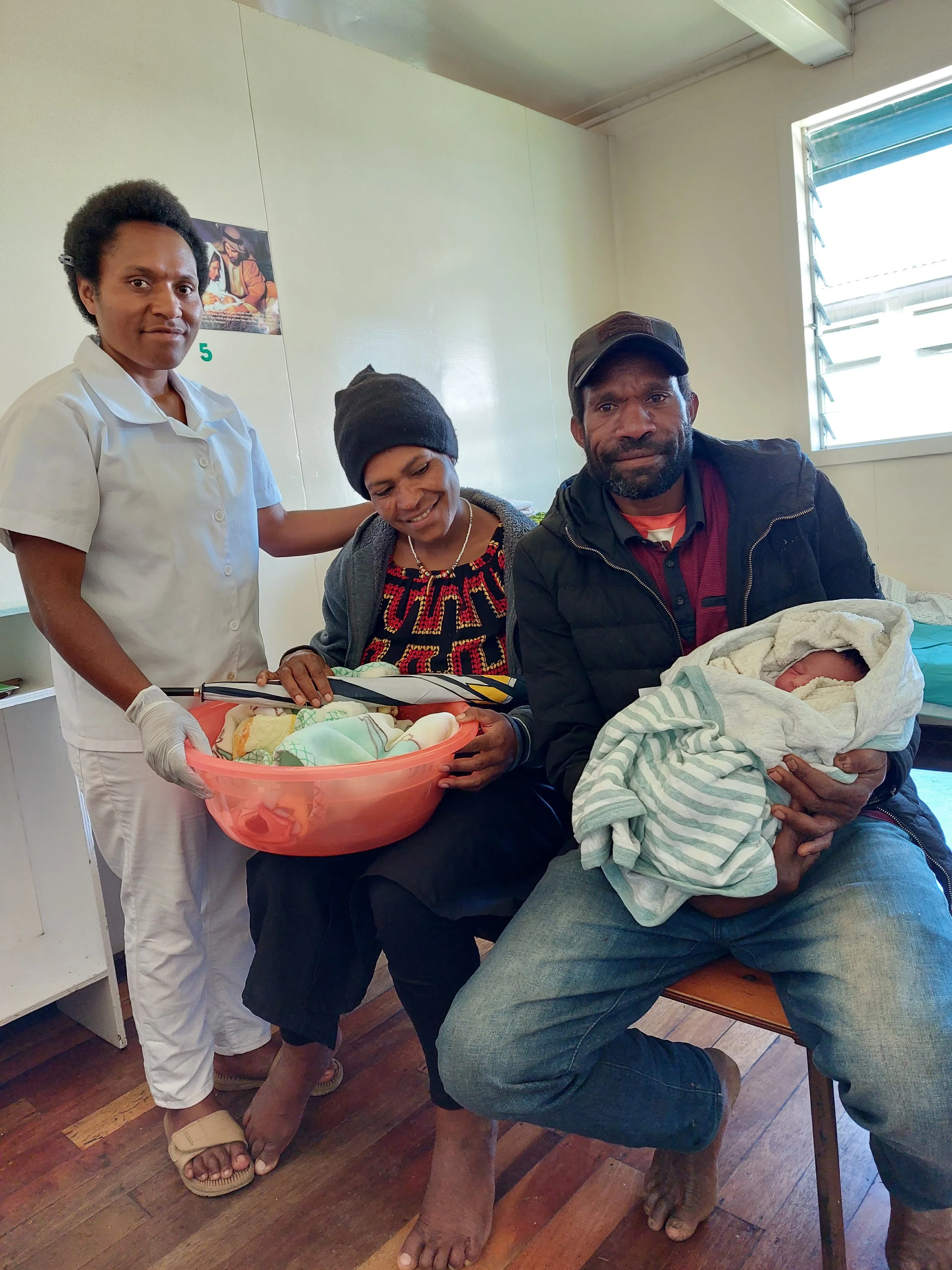 A group of four people, including a nurse, a woman, and two men, with a newborn baby wrapped in a blanket. The nurse is holding a bowl with baby clothes, and one man holds the newborn, all inside a room with a window.