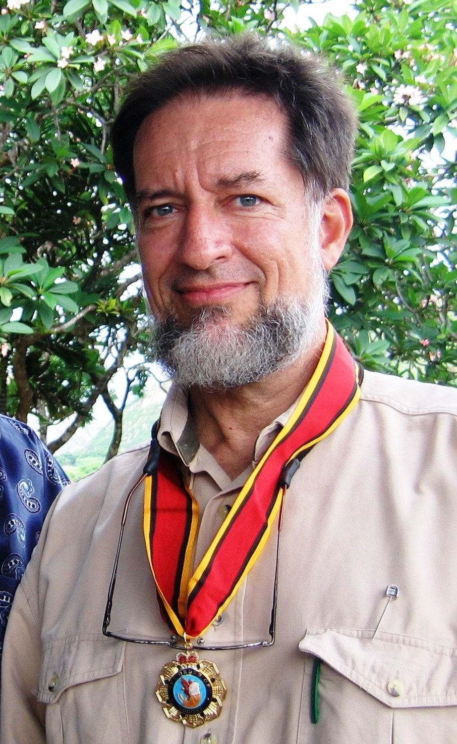A middle-aged man with a beard and mustache, wearing a beige uniform shirt and a medal around his neck, standing outdoors in front of green leafy trees.