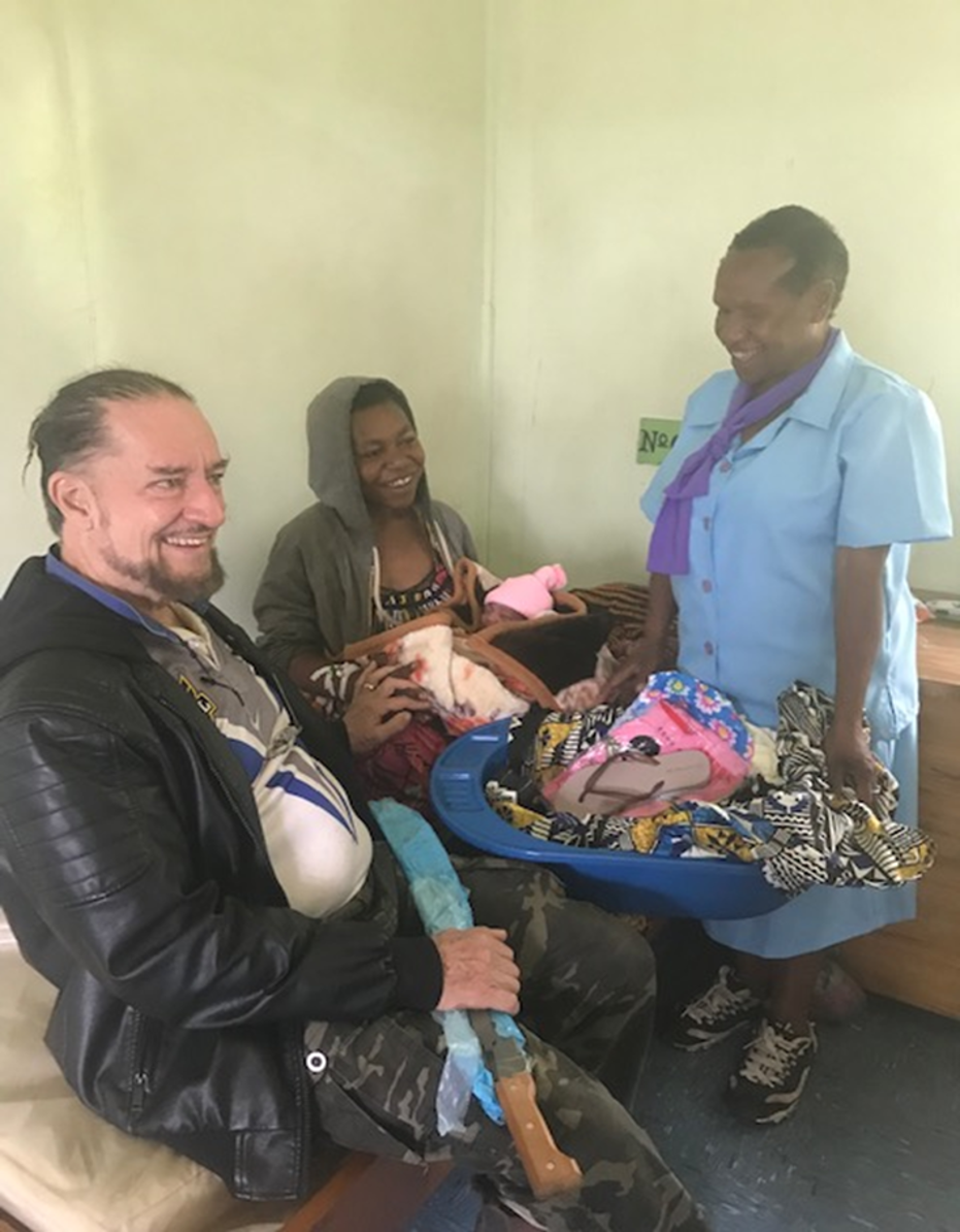 A diverse group of three adults, two men and one woman, inside a room, with the woman holding a newborn baby wrapped in pink. The woman is smiling and sitting on a bench, while the man in the leather jacket sitting next to her is smiling with a leg cast. The third person, a woman in a blue uniform, is standing and holding a basket of colorful clothes and garments.