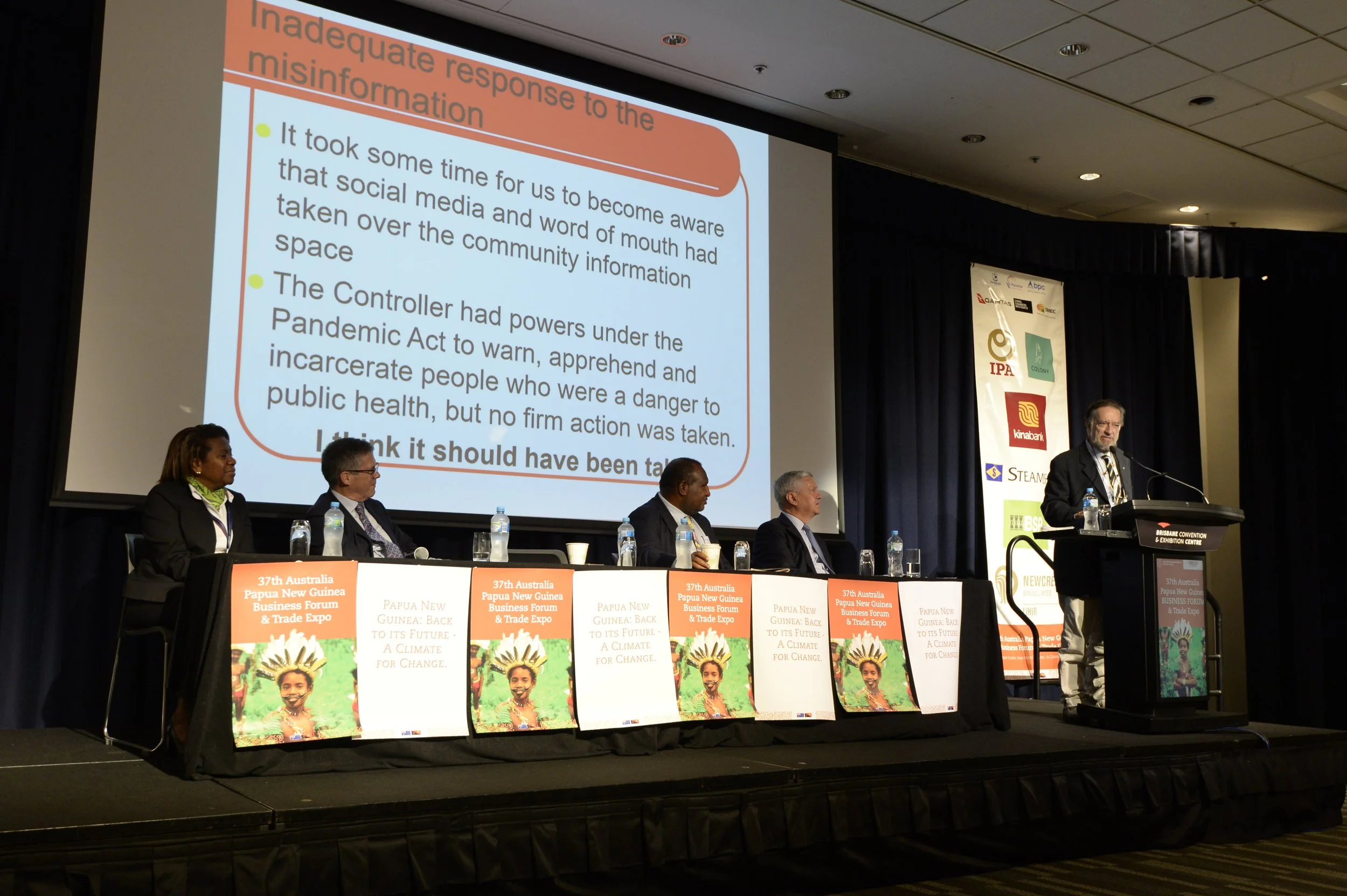 A panel of five people sitting at a table during a conference or forum, with a large presentation screen behind them displaying a slide. The slide contains text about misinformation and social response, with bullet points. The table has banners with information about the Papua New Guinea Business Forum & Trade Expo, featuring images of a young girl with a bright smile and traditional face paint. One person is standing at a podium speaking. The background has a curtain with logos and signs for the event.