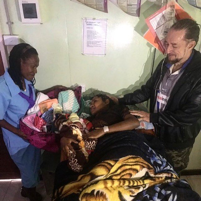 A woman lying in a hospital bed surrounded by medical staff and visitors, with a basket of supplies nearby.