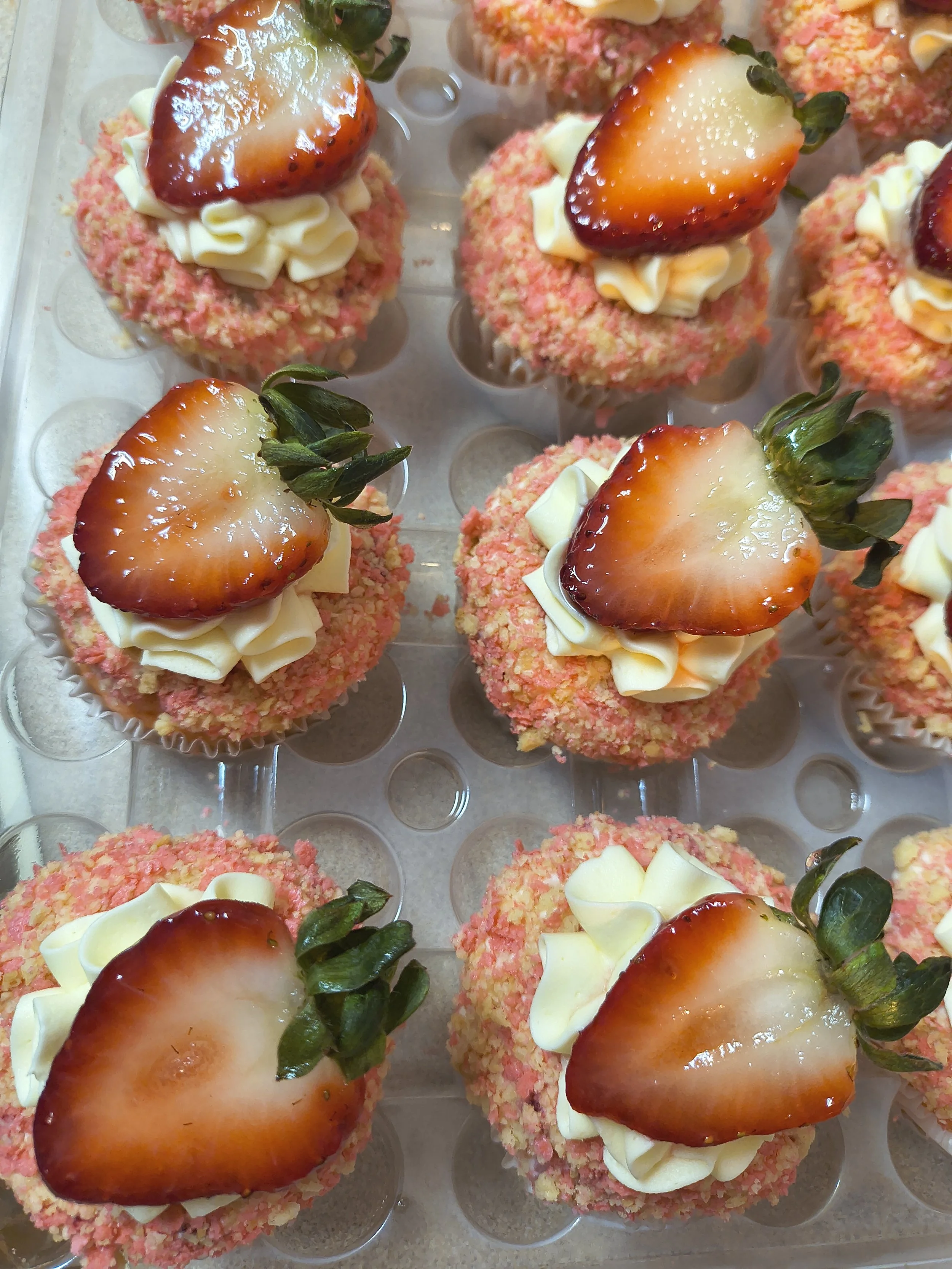 Frosted cupcakes topped with halved strawberries and white chocolate curls on a clear plastic tray.