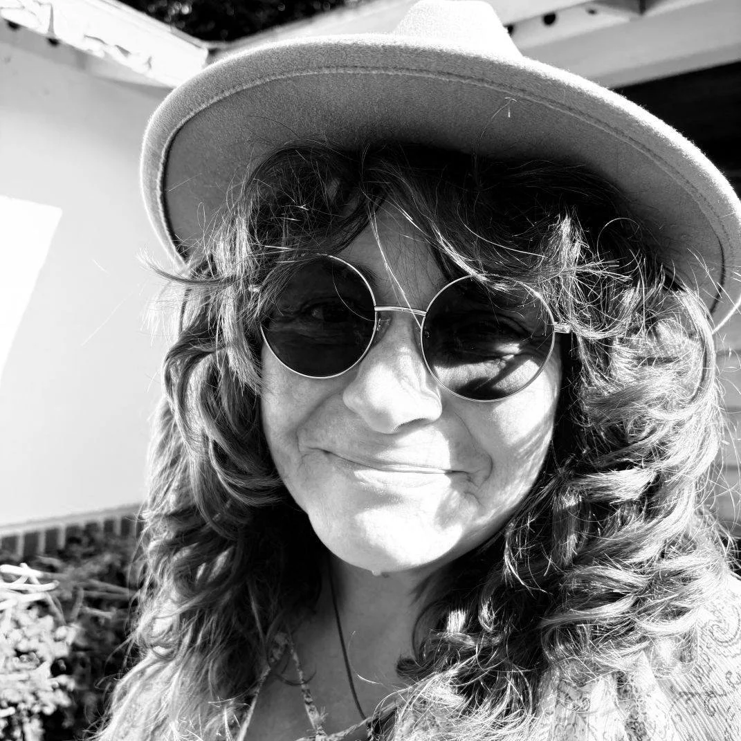 Woman with curly hair wearing sunglasses and a wide-brimmed hat, outdoors, smiling.