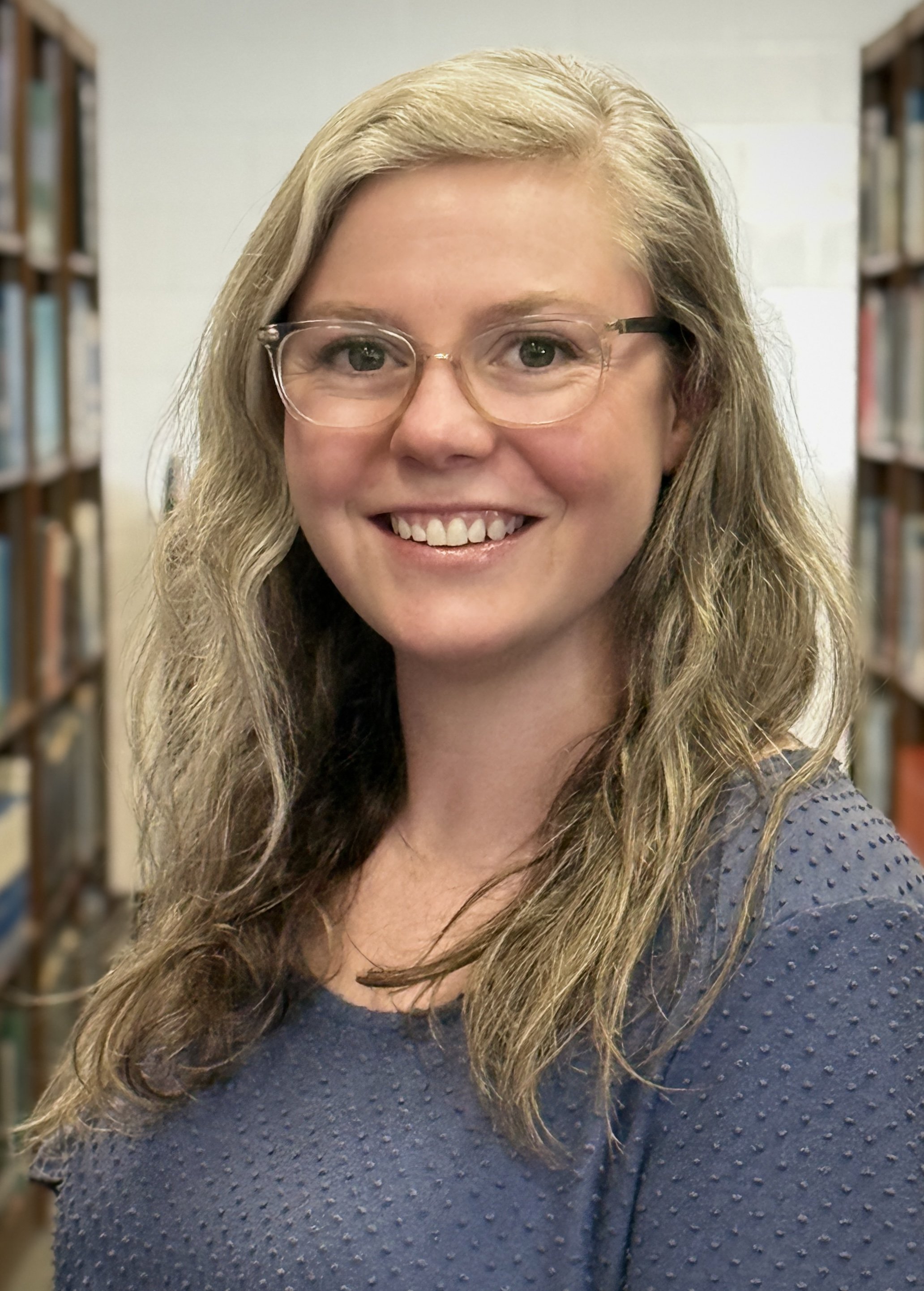 A woman smiling with glasses in a library.