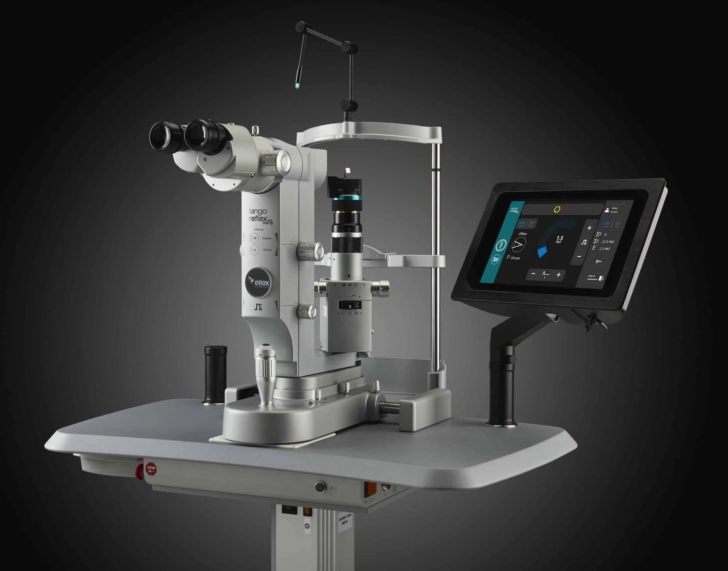 A slit lamp microscope used for eye examination, with a digital display screen beside it, placed on a gray table against a dark background.