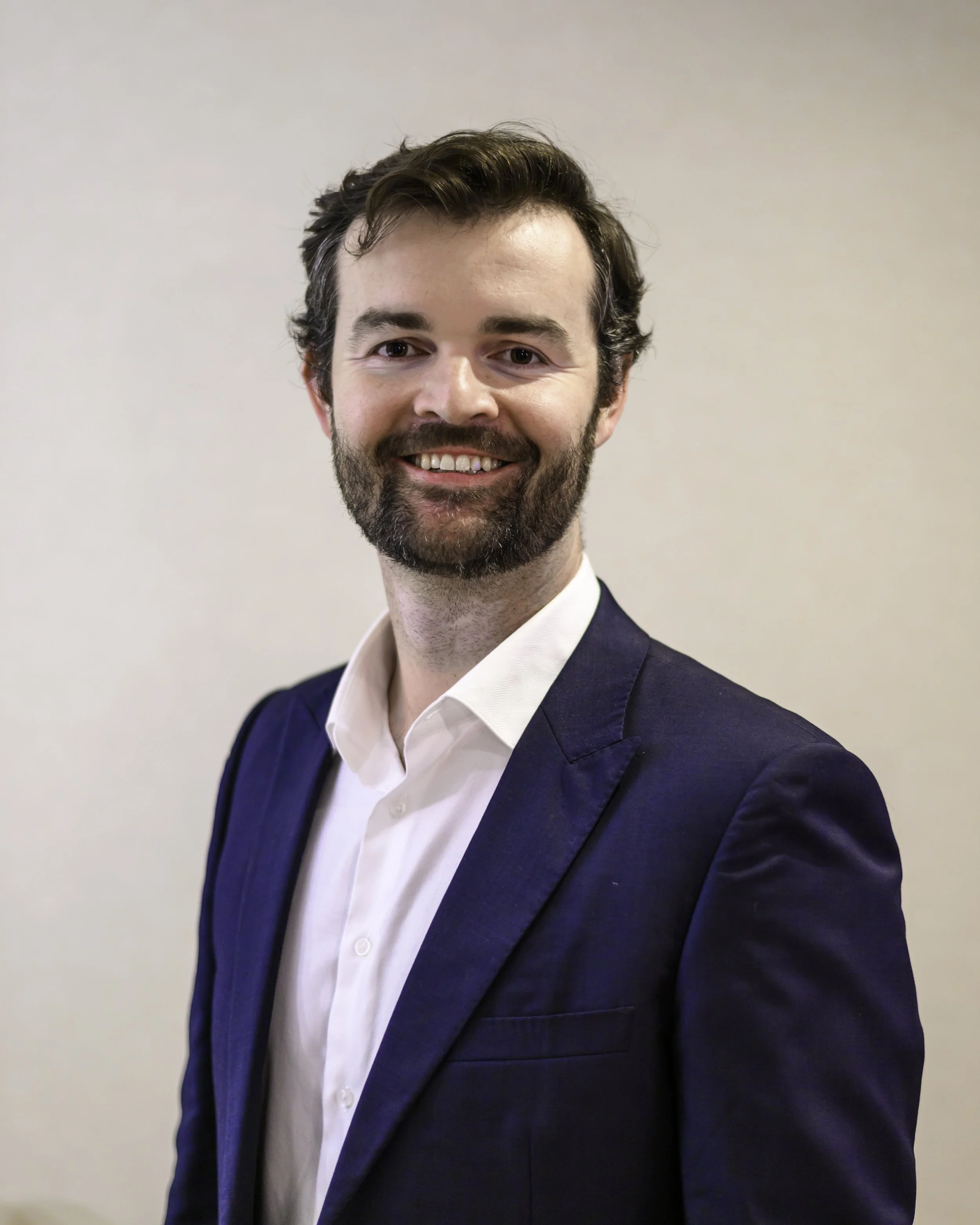 A smiling man with dark brown hair and a beard, wearing a navy blue suit jacket over a white dress shirt, standing against a plain light-colored background.