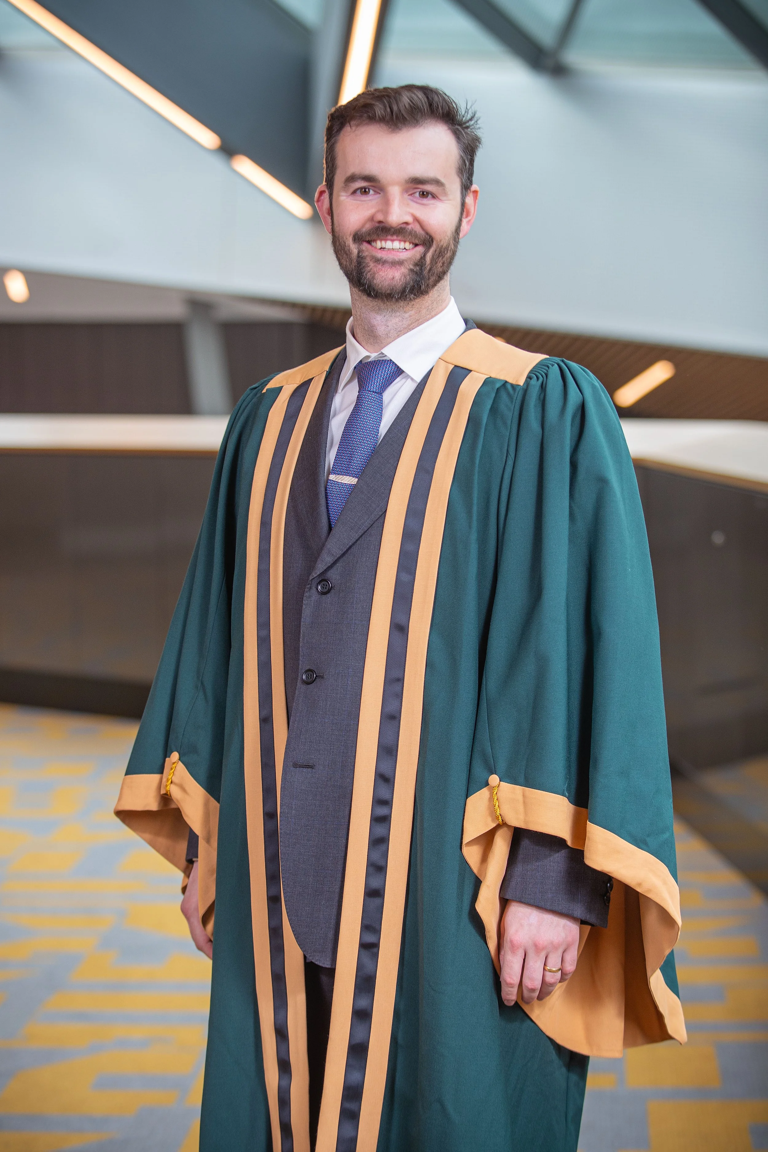 A smiling man in a graduation gown and cap standing in a modern building with colorful flooring and a high ceiling.