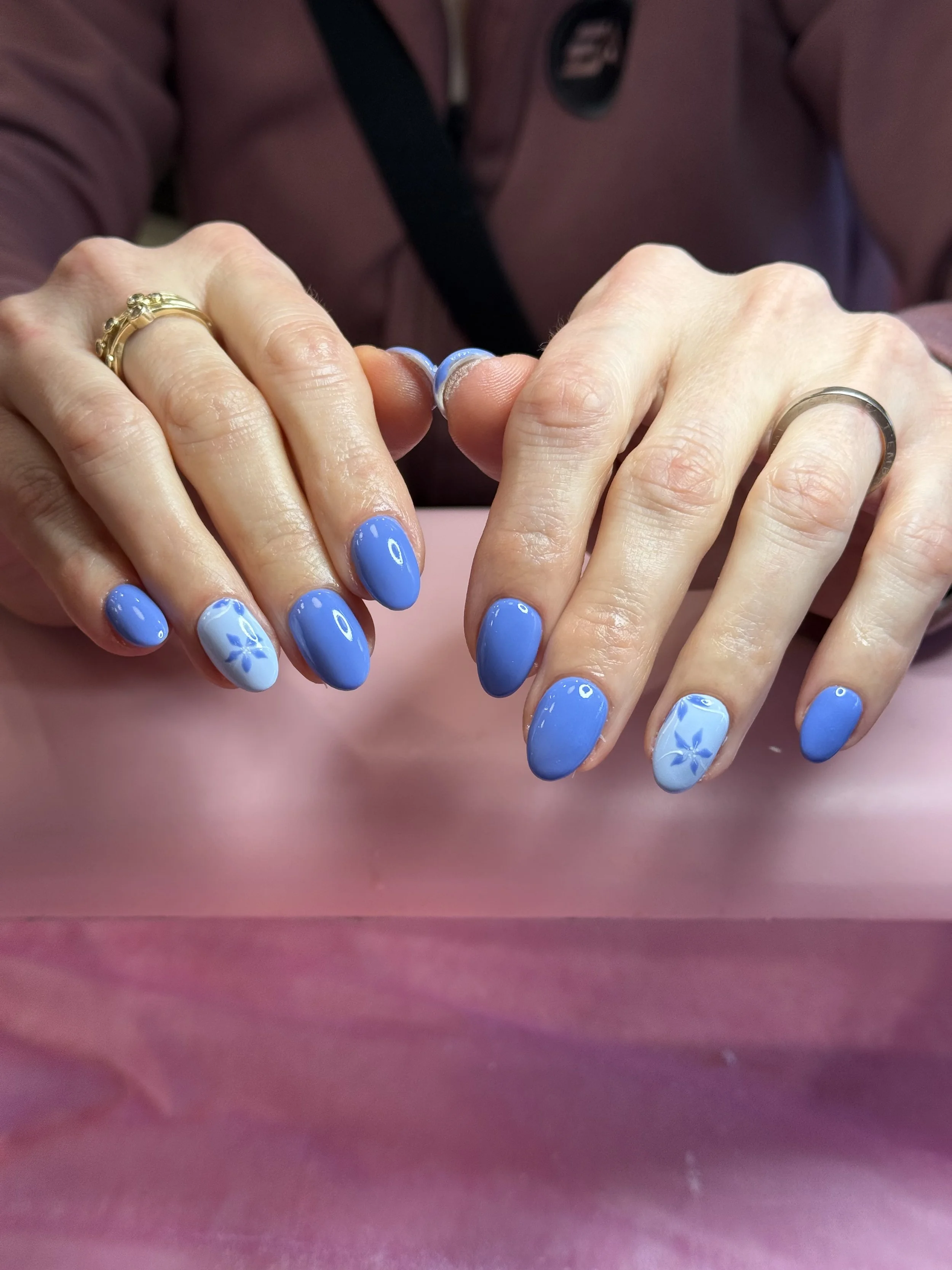 Close-up of hands with blue painted nails, wearing rings, resting on a pink surface, with a person's torso and outfit partially visible in the background.