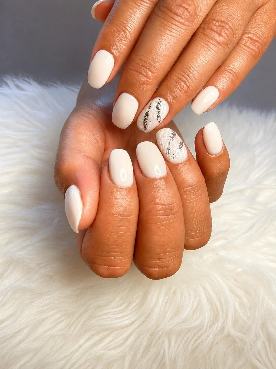 Close-up of manicured hands with white and silver nail designs, resting on a soft, white furry surface.