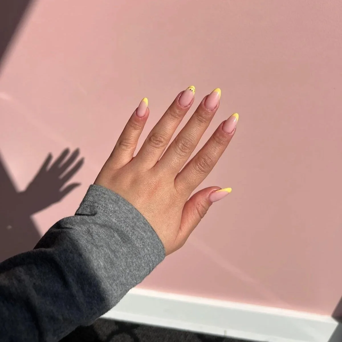 A person's left hand with yellow French tips, palm down against a pink background, casting a shadow.