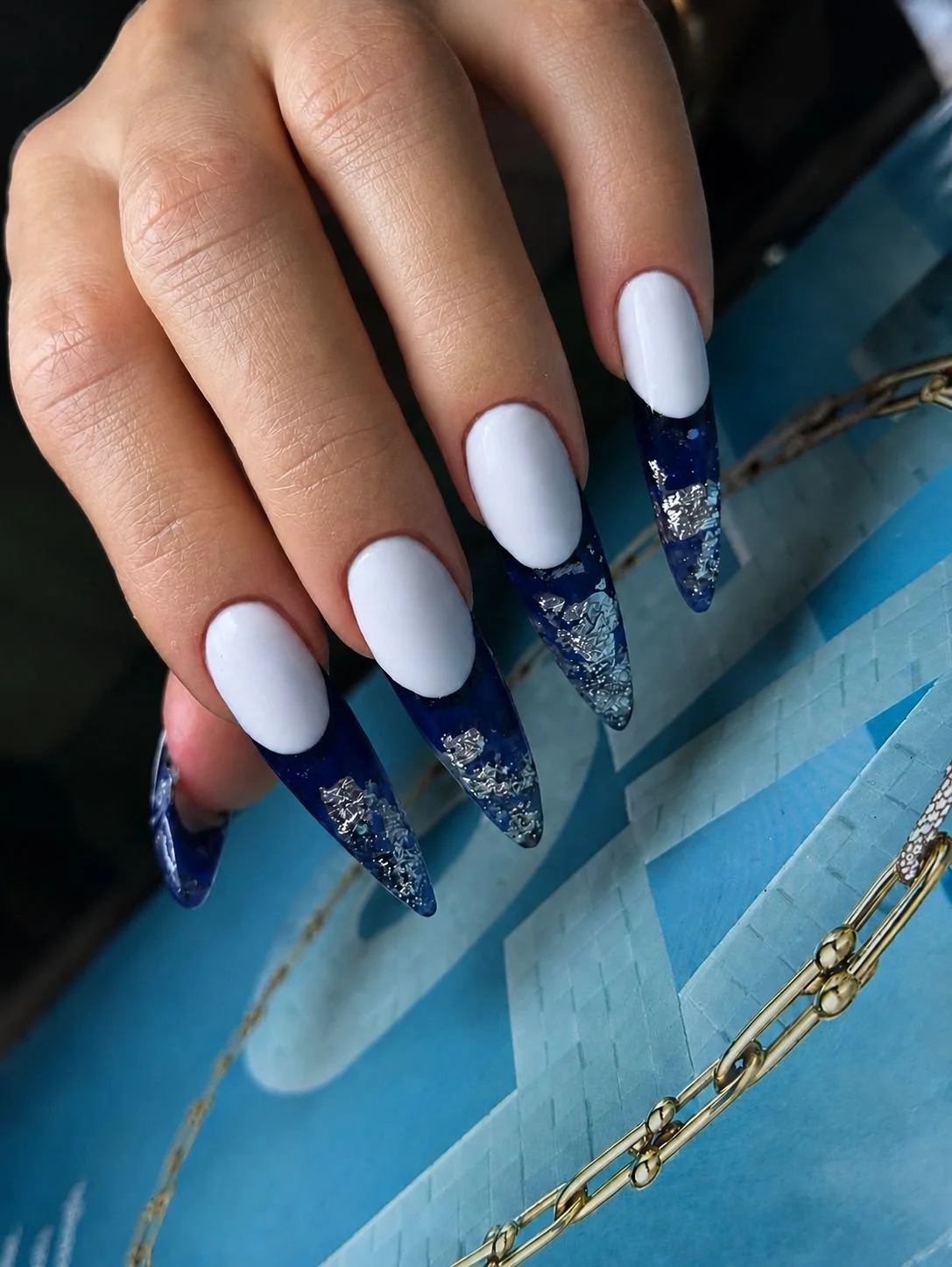 Close-up of a hand with long, almond-shaped nails painted white with dark blue tips and silver foil accents.