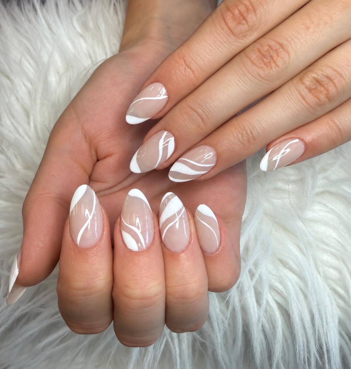 Close-up of a woman's hands with elegant French manicure featuring white swirling lines on a clear base, resting on a soft white furry surface.