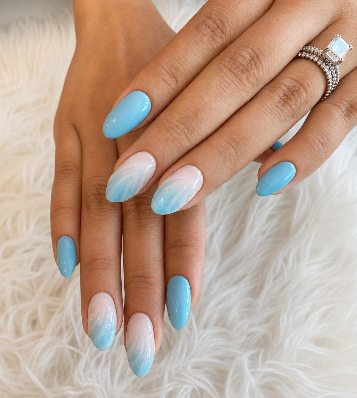 Close-up of a woman's hands with light blue almond-shaped nails, some with white and blue marbled designs, resting on a fluffy white surface. She wears a large square-cut gemstone ring on her ring finger.