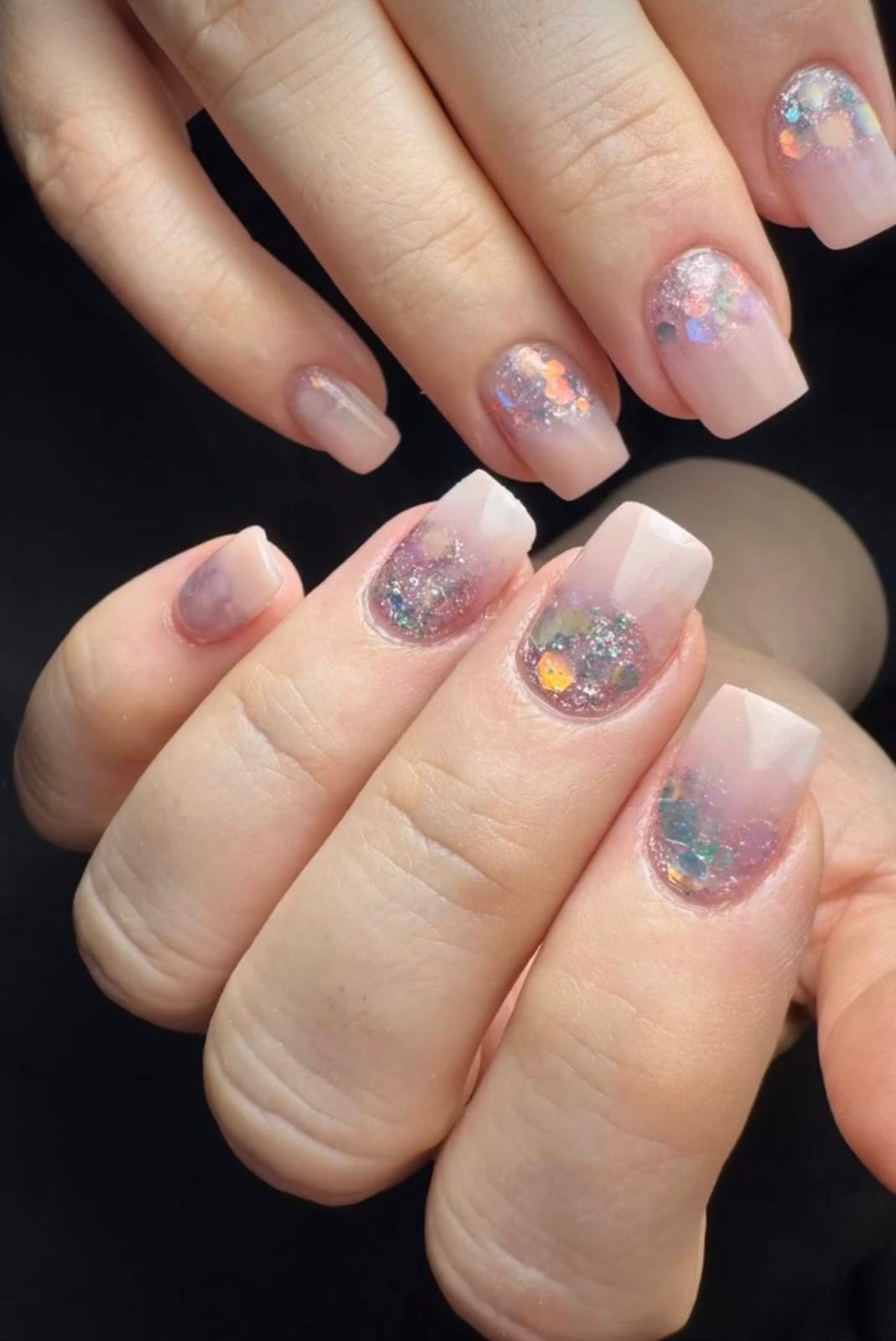 Close-up of a person's hands displaying manicured nails with a clear base and colorful, iridescent glitter.
