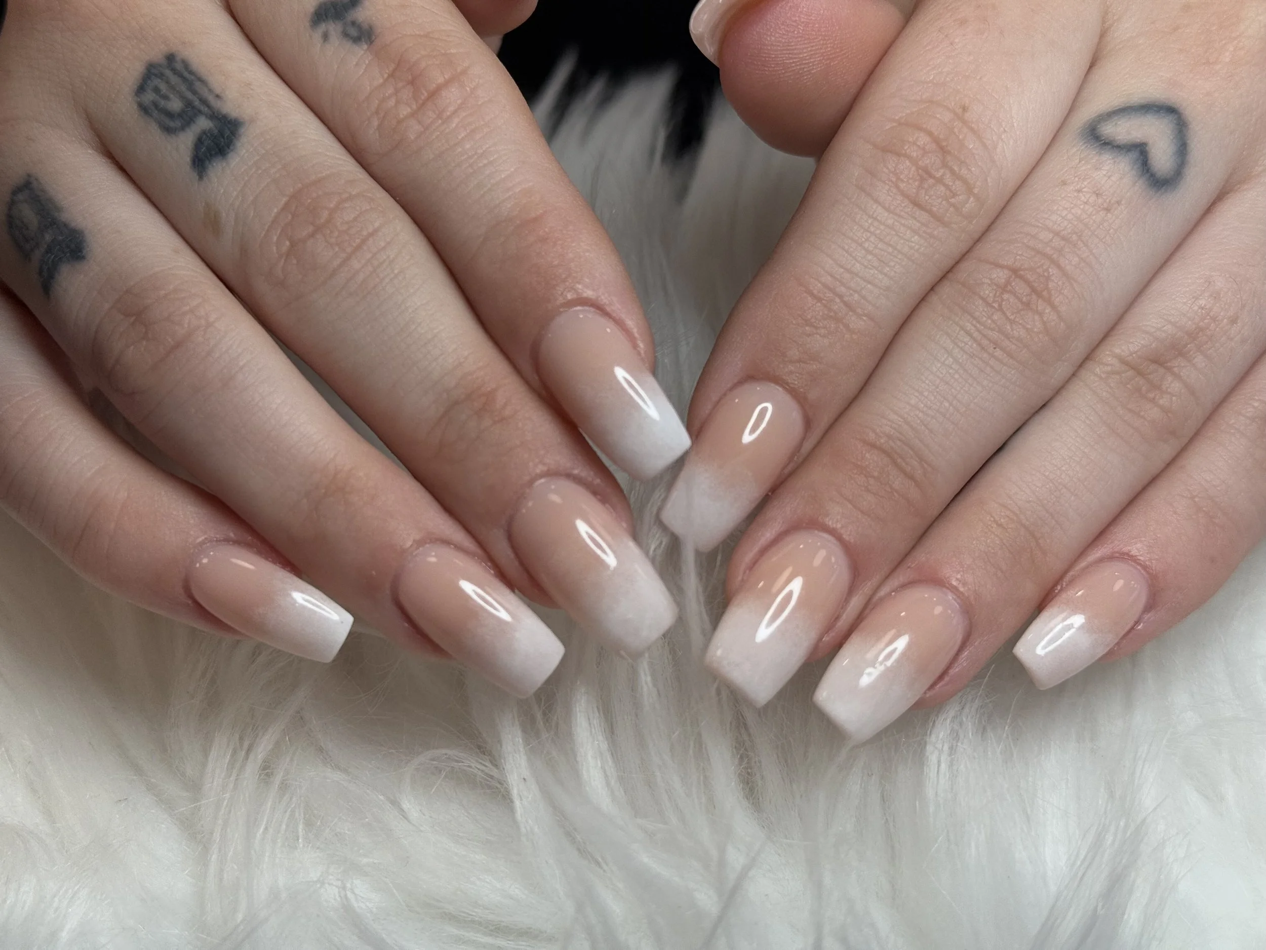 Close-up of a person's hands with tattoos, finger nails painted with nude-colored polish, resting on a white furry surface.