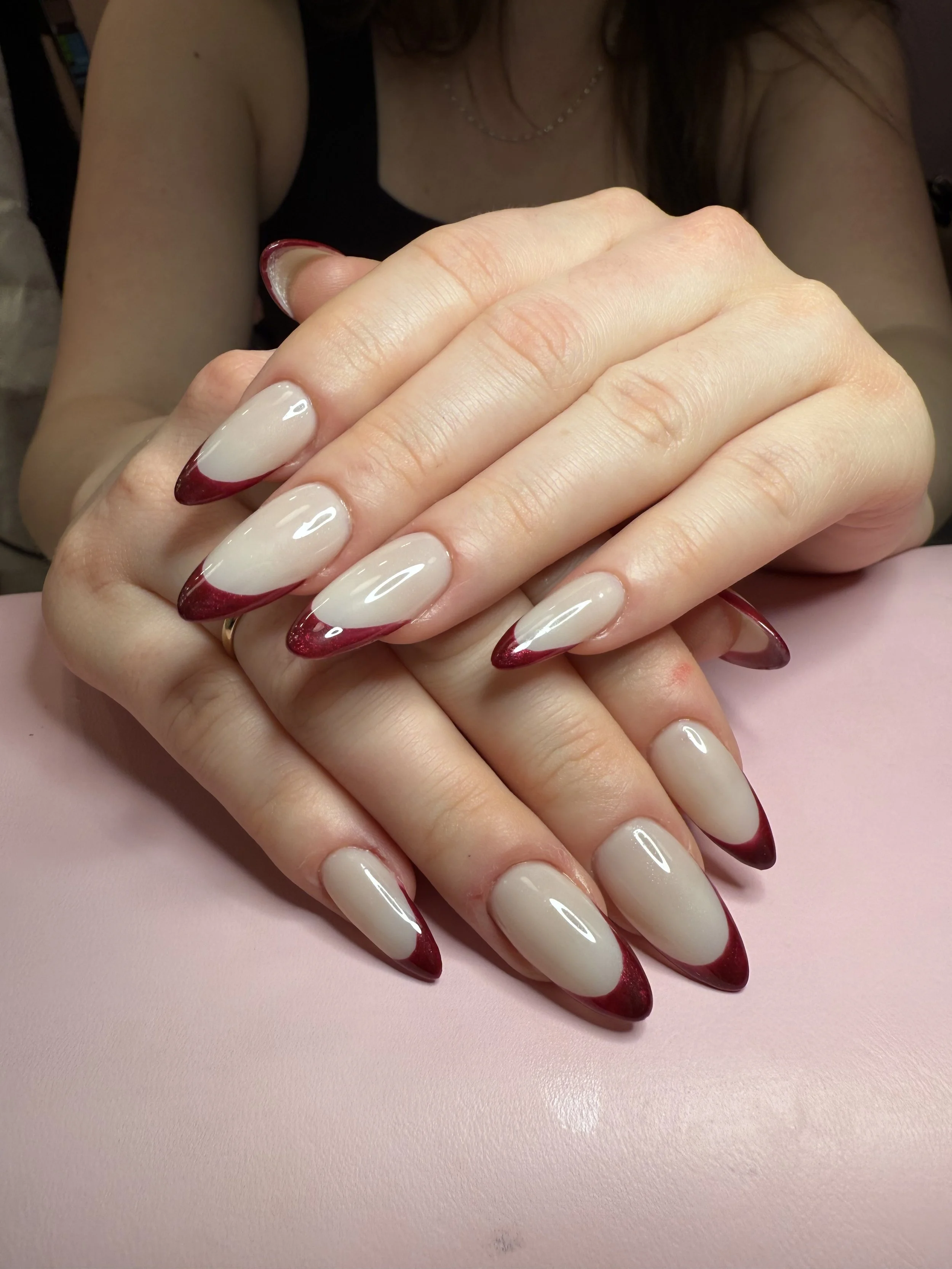 Close-up of a woman's hands with long, almond-shaped nails that are painted with a glossy beige color and have red tips. The woman is wearing a black sleeveless top and a delicate necklace, and her hair is brown and slightly wavy. The background incl