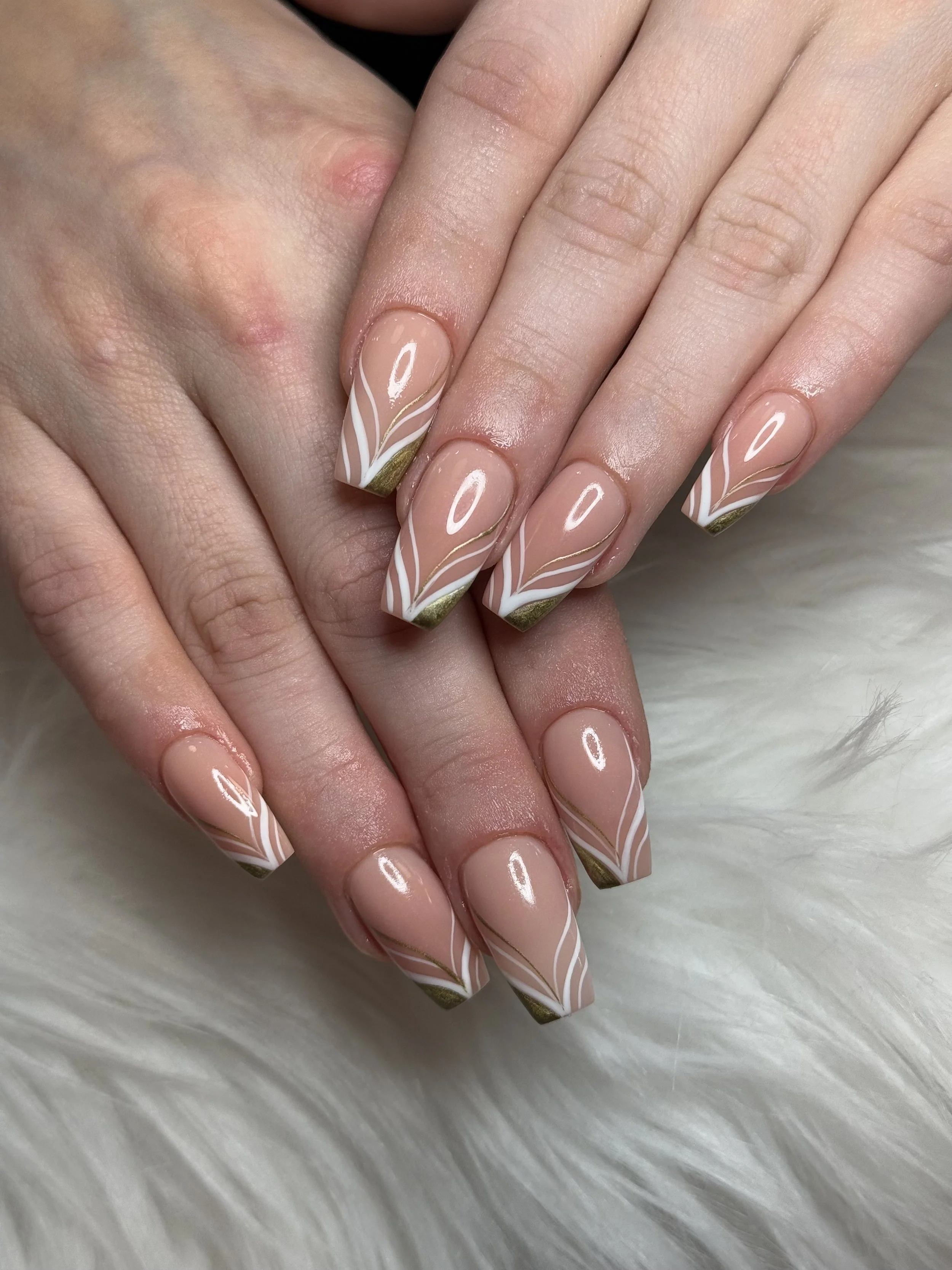 Close-up of manicured nails with a nude base color featuring white and gold wavy line designs and gold tips, resting on a soft white furry surface.