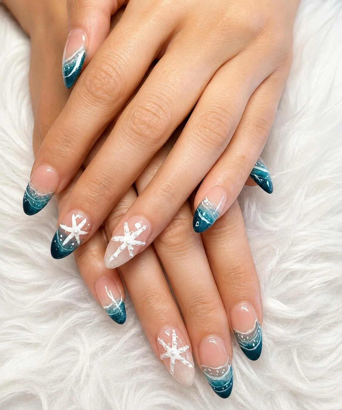 Close-up of hands with decorated French manicured nails featuring teal, white, and glitter accents and snowflake and starfish designs, resting on a white furry surface.