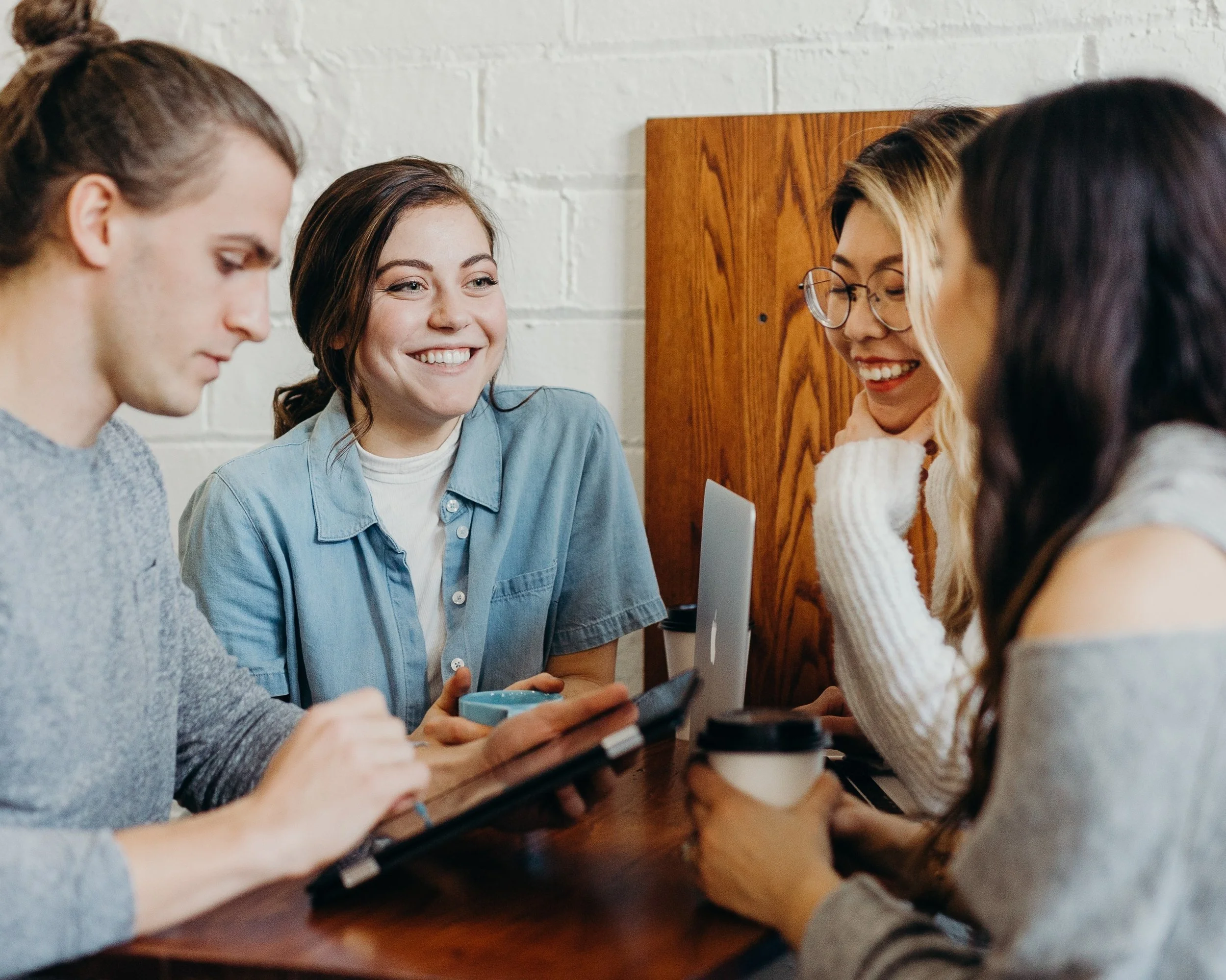 Four young women sitting at a table in a cafe, engaged in conversation. One is using a tablet, another holding a coffee cup, and the conversation appears friendly and lively.