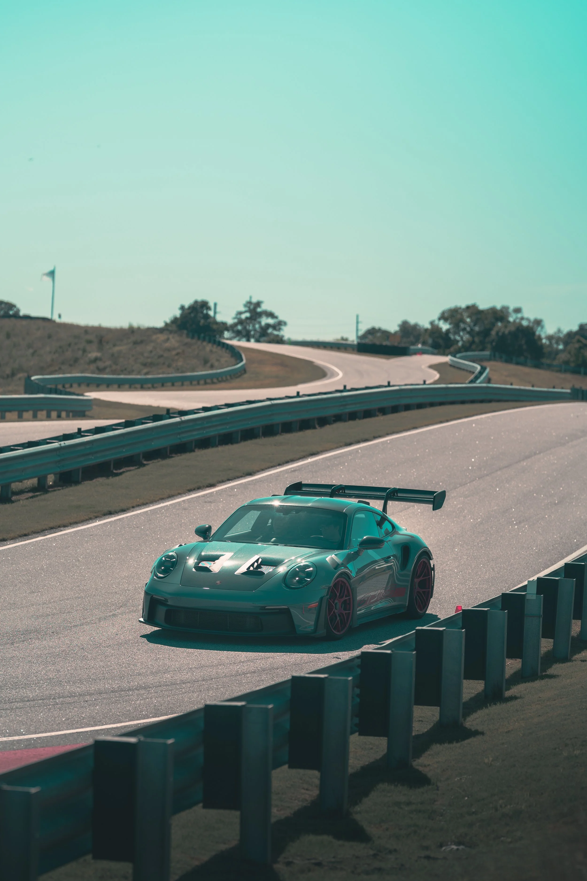 A black sports car with a large rear wing driving on a winding race track with guardrails and grassy hills, under a clear sky.