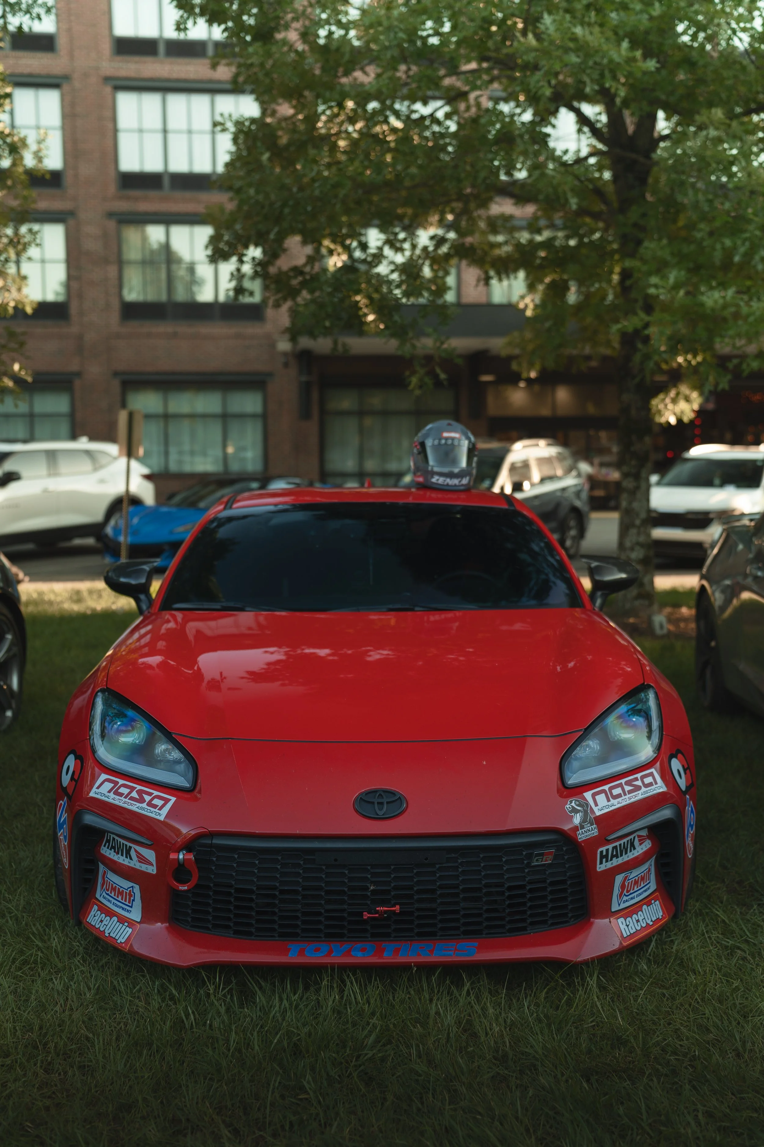 Red race car with stickers, parked on grass, with a bike helmet on its roof, in a parking lot with trees and multi-story brick building in the background.