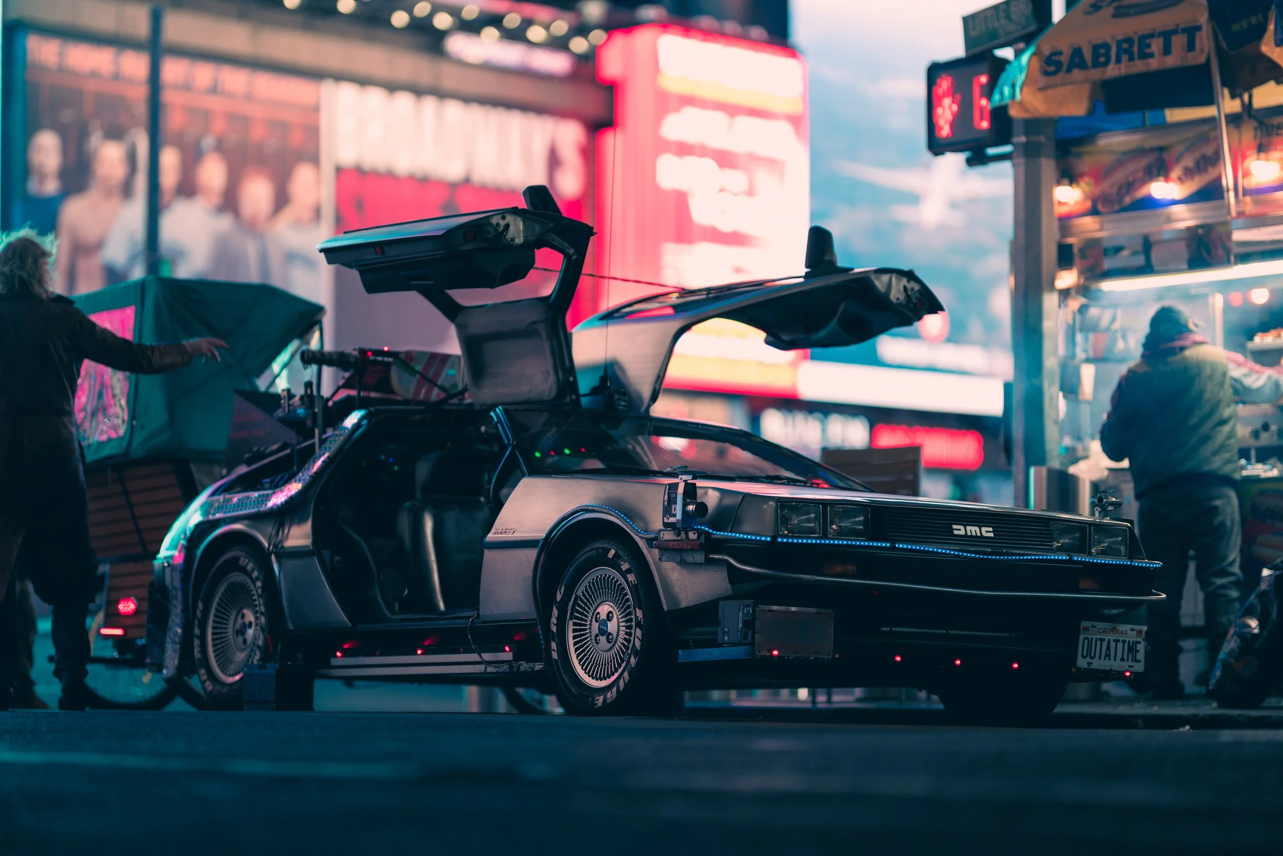The image shows a scene from a sci-fi movie or a car display setup featuring the DeLorean time machine from 'Back to the Future.' The DeLorean is parked on a flatbed with its gull-wing doors open and is illuminated with colorful lights. The background includes neon signs and digital billboards, creating a vibrant, urban night-time atmosphere.