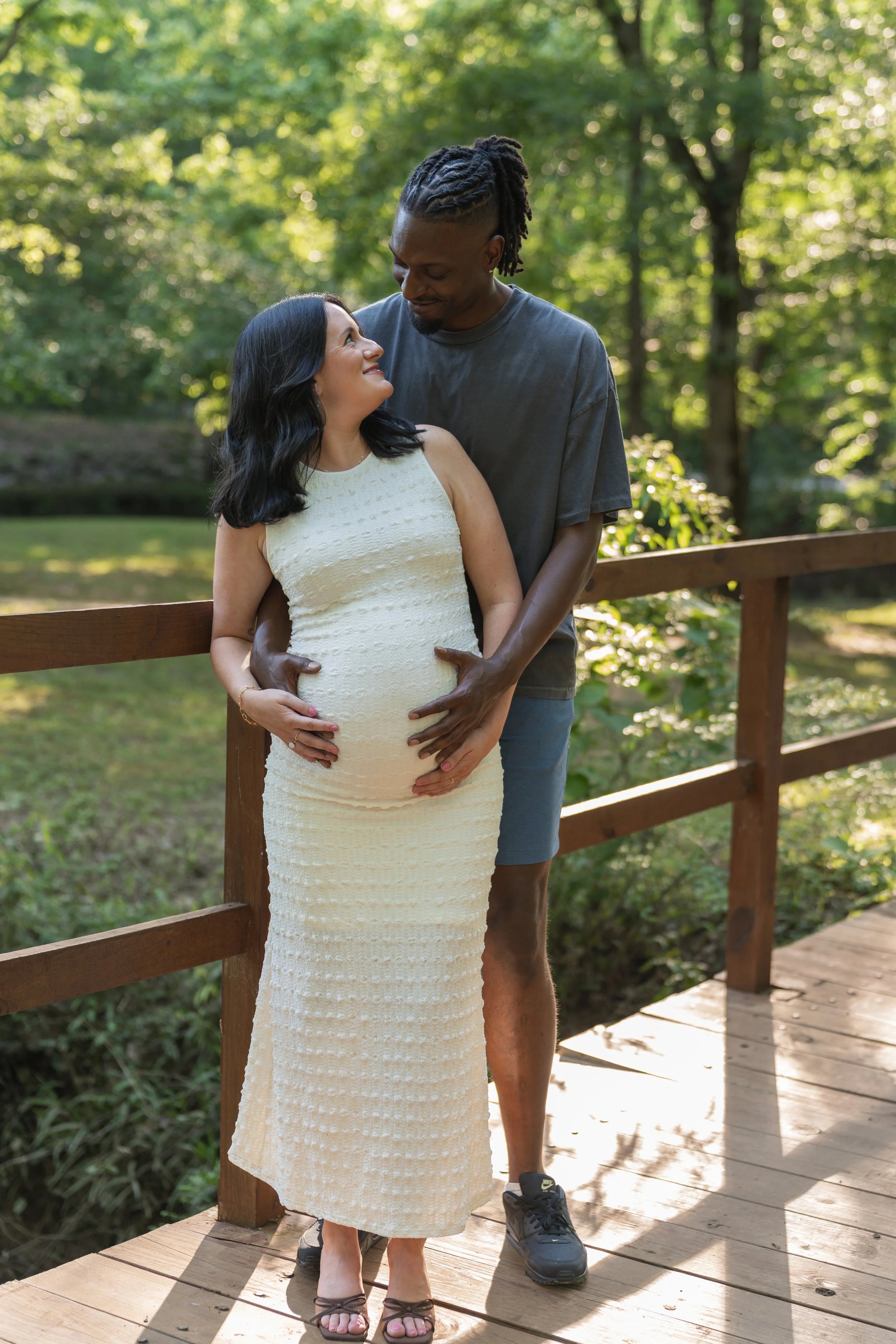 A pregnant woman in a white dress and sandals standing on a wooden bridge, looking lovingly at a tall man with dreadlocks dressed in a gray T-shirt, behind her with his hands on her belly, in a green forest setting.