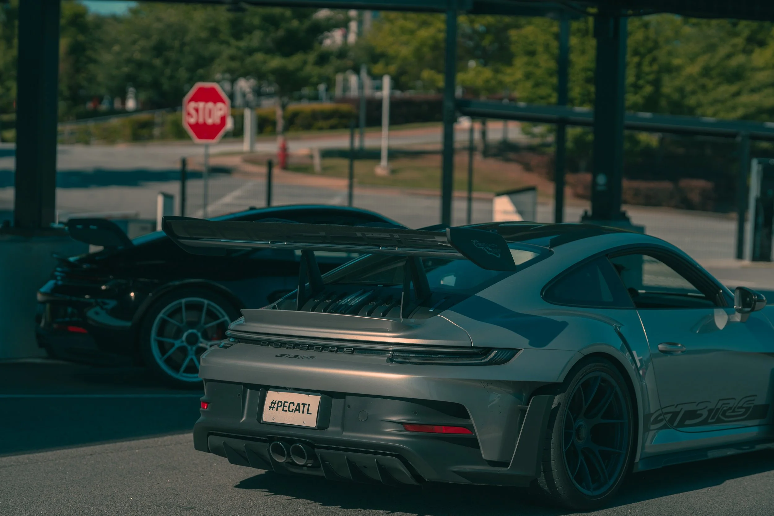 Rear view of a silver Porsche 911 sports car with a spoiler. A black sports car is parked next to it. In the background, a stop sign and trees are visible.